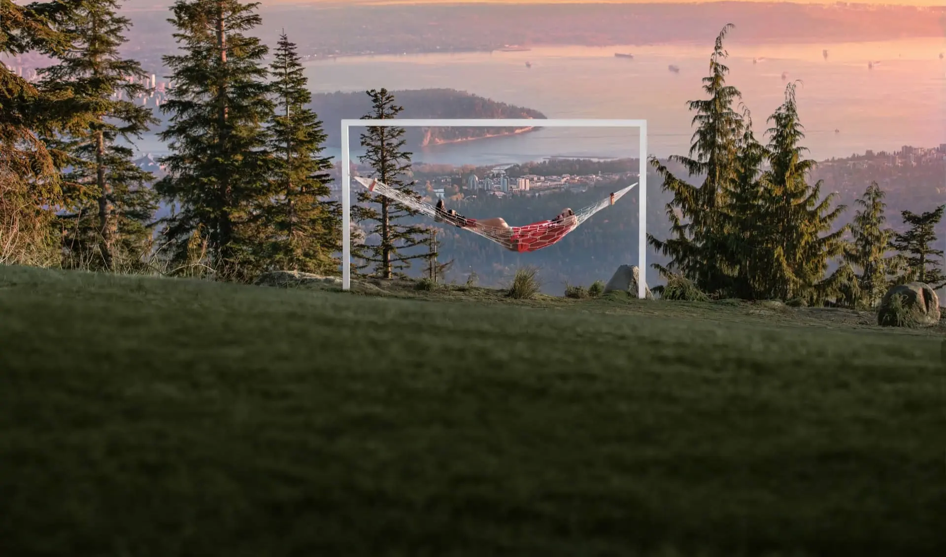 Person lying in a red hammock suspended within a white frame on a grassy hill with trees and a city and water view at sunset.
