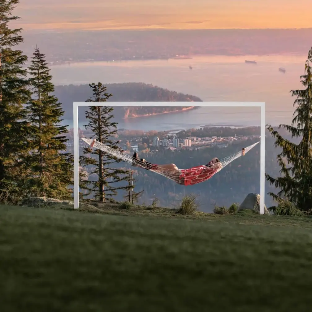 Person lying in a red hammock suspended within a white frame on a grassy hill with trees and a city and water view at sunset.