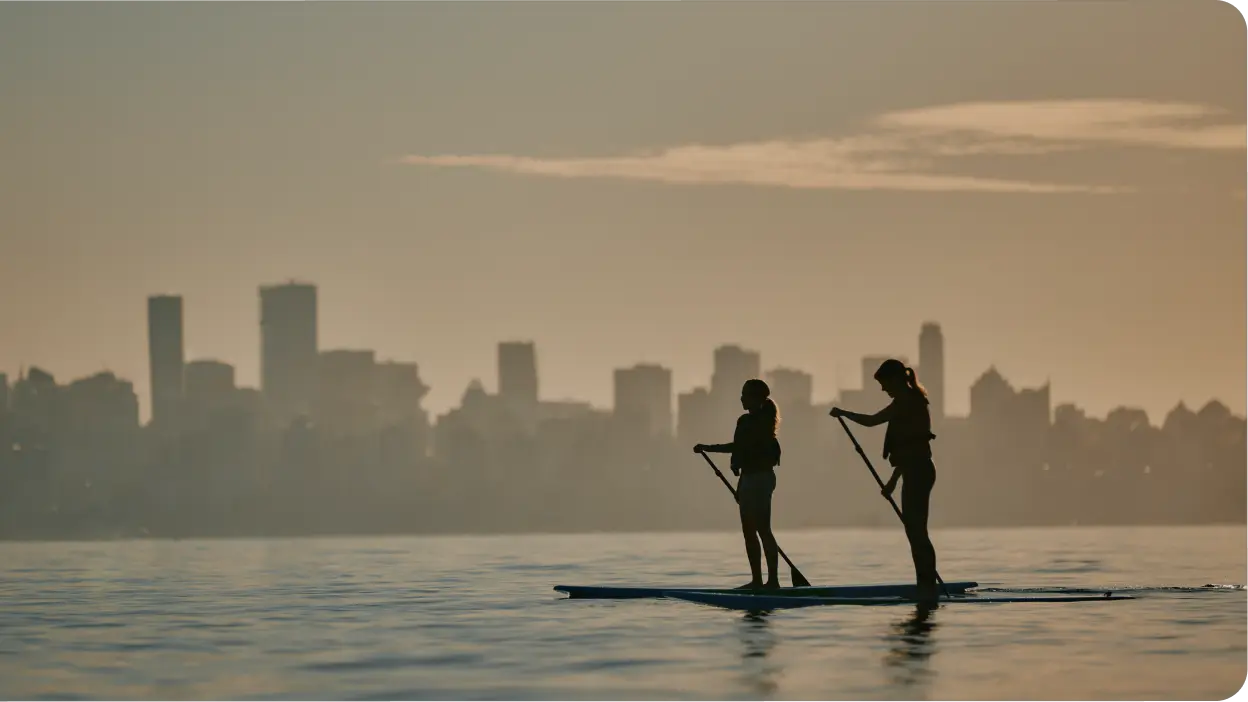 Two people paddleboarding on calm water at sunrise or sunset, with a city skyline silhouetted in the background under a partly cloudy sky—a perfect addition to your Vancouver itinerary.