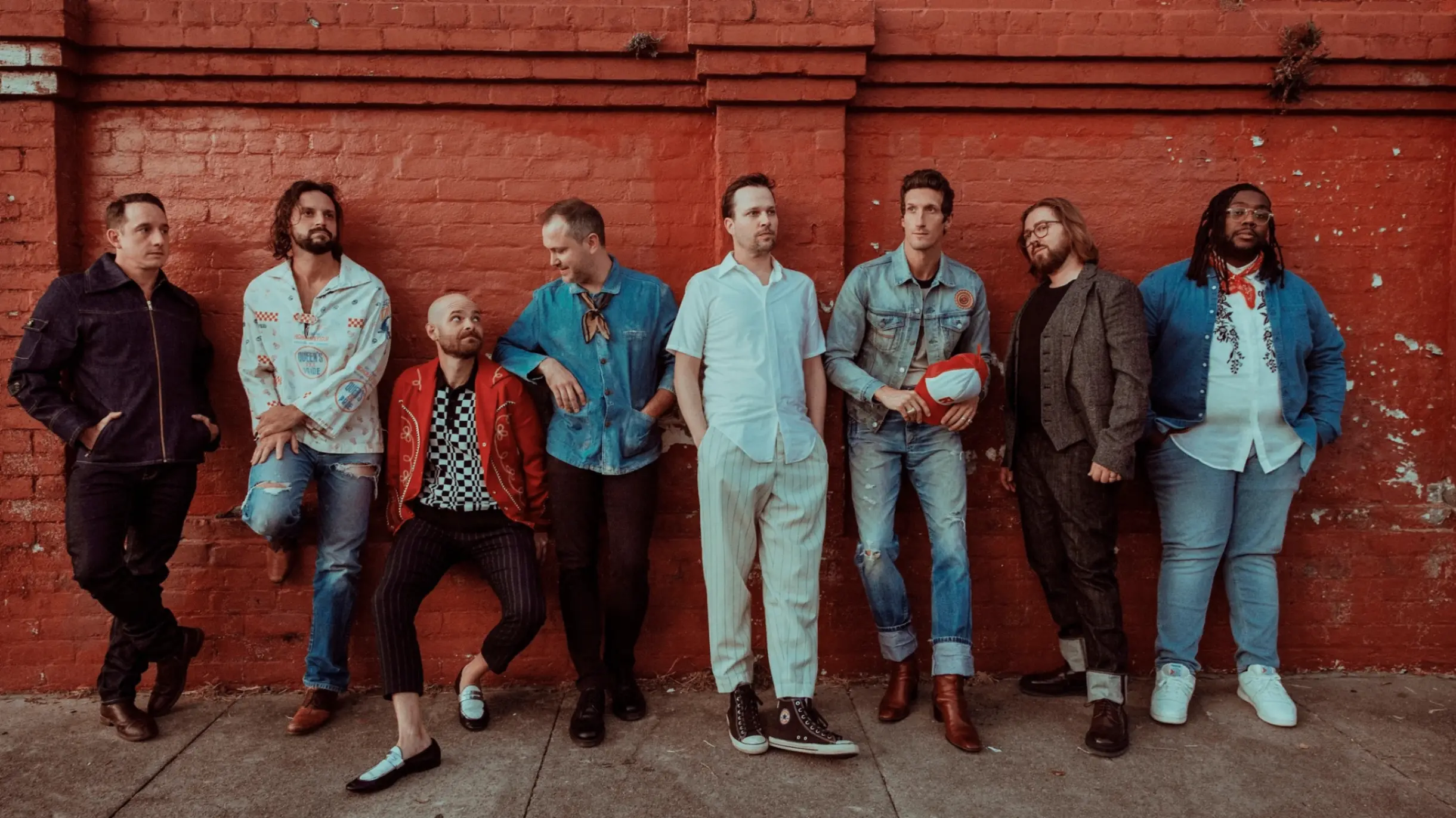 Eight people standing and leaning against a red brick wall, dressed in casual and semi-formal clothing.