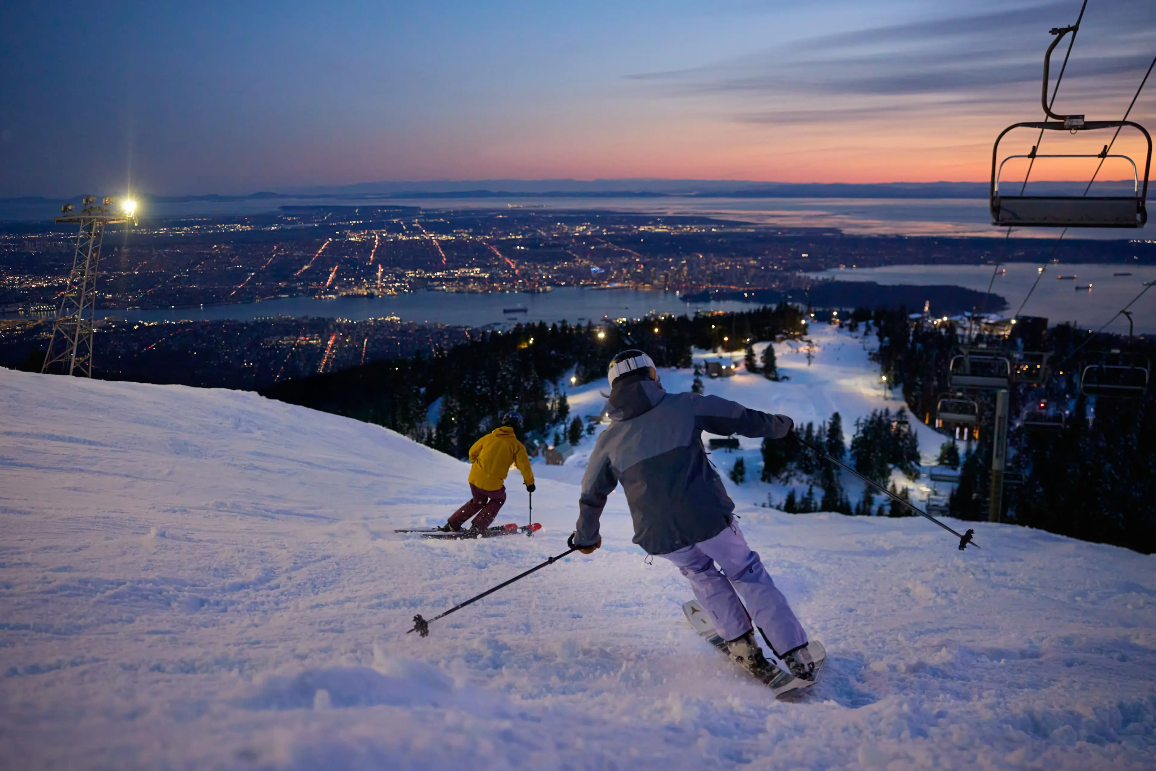 Two skiers descending a snowy slope at dusk with city lights and a chairlift in the background.