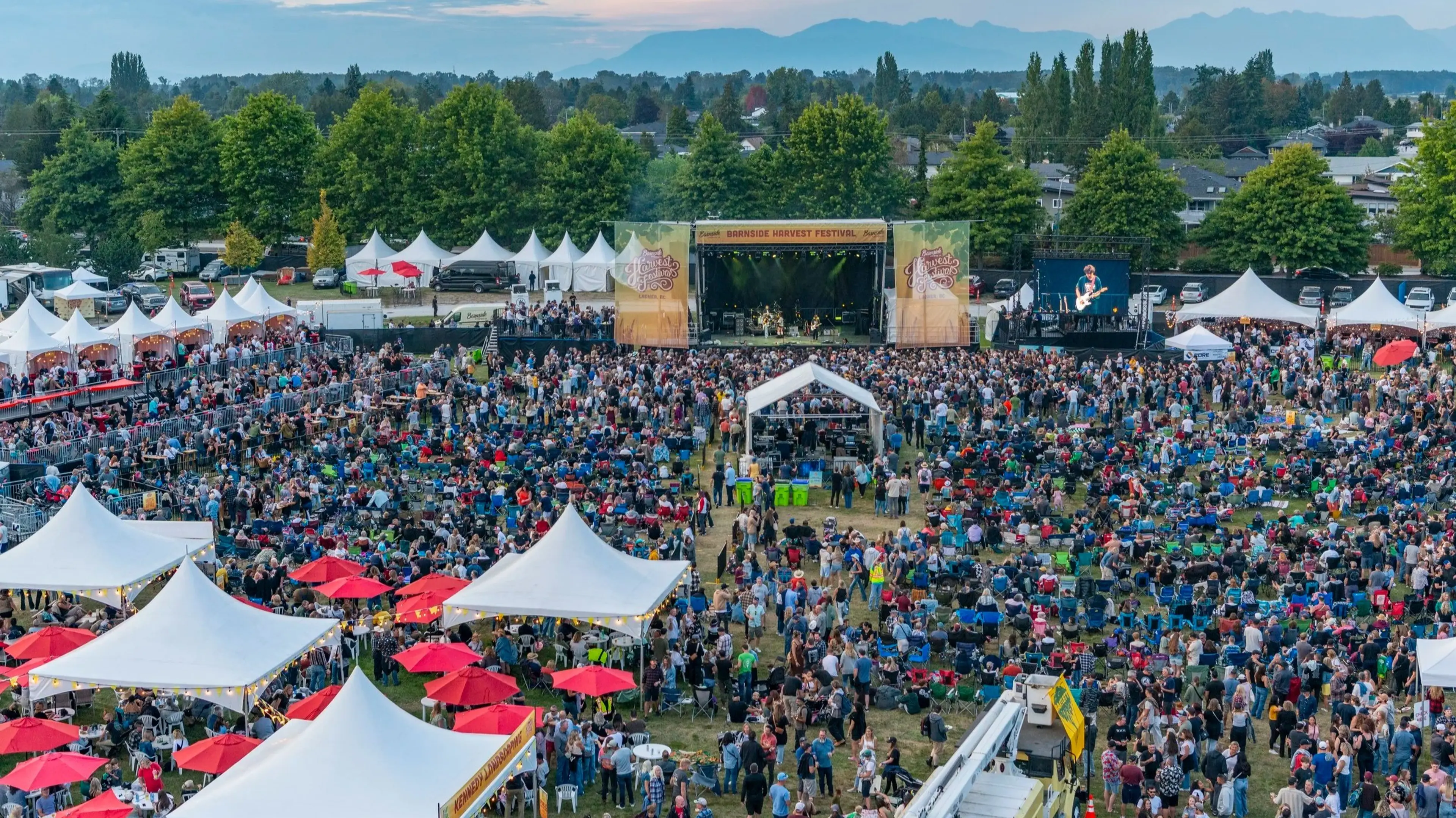 Large outdoor music festival crowd with white tents, red umbrellas, and a stage with a band performing.
