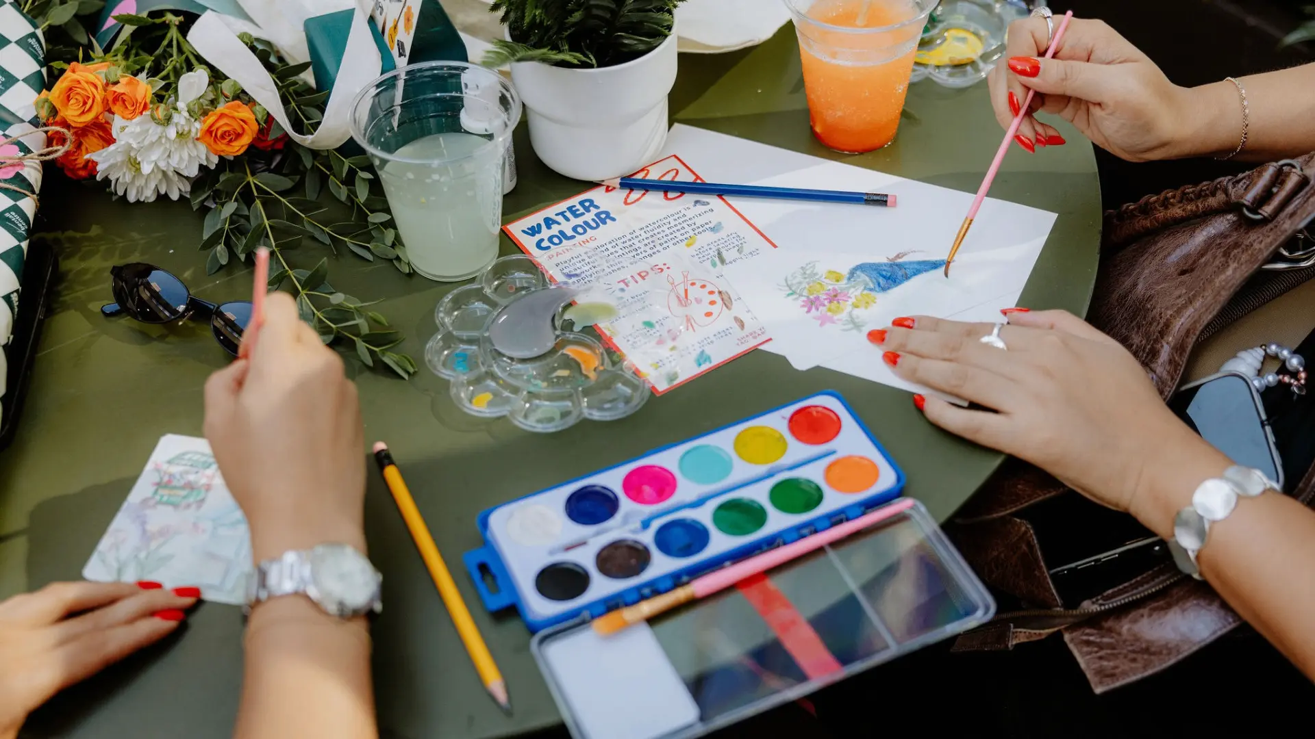 Two people painting with watercolors on paper at a table with flowers, drinks, and art supplies.