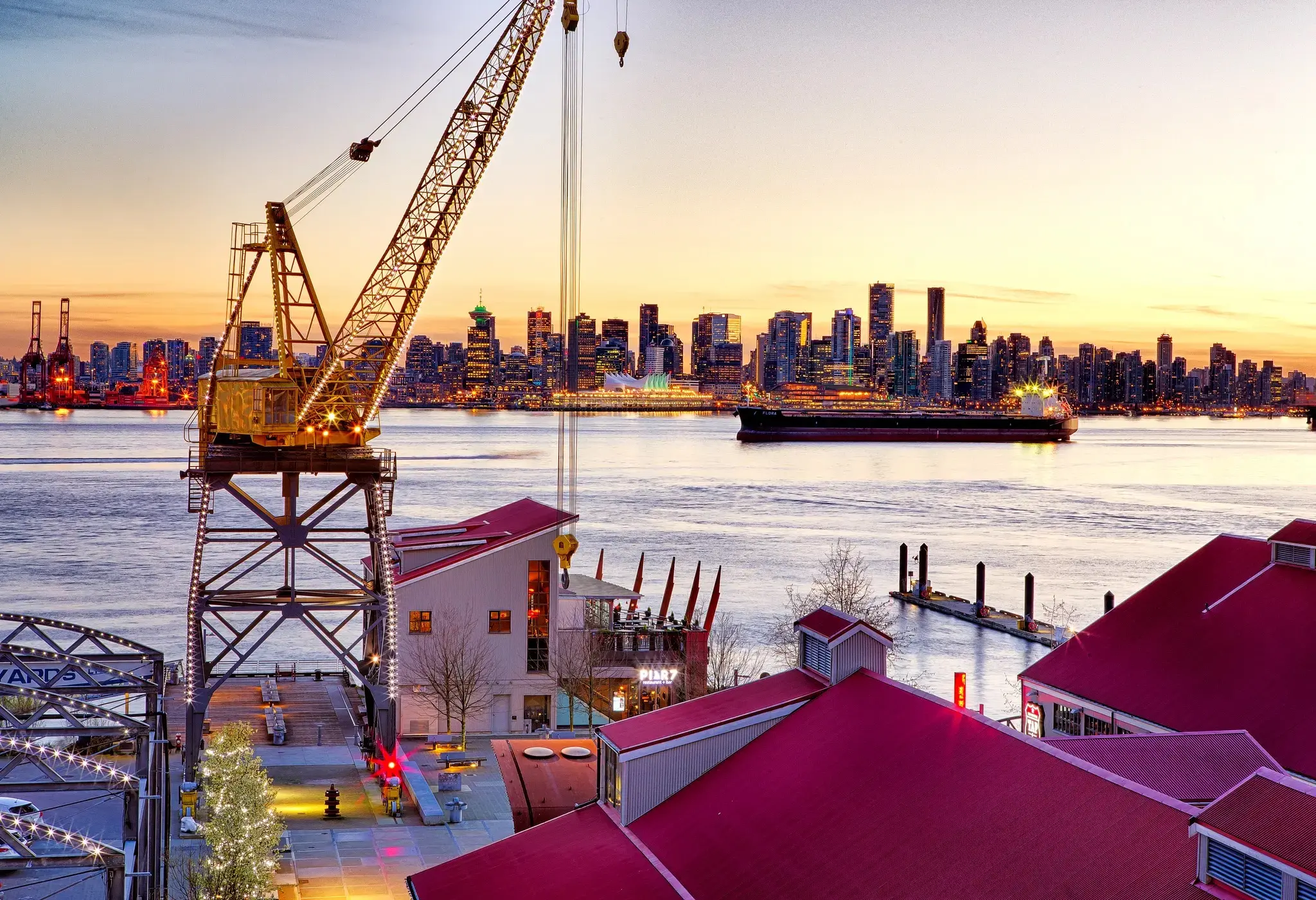 Lower Lonsdale dock at sunset with a crane, a barge in the ocean, and skyscrapers against a sunrise.