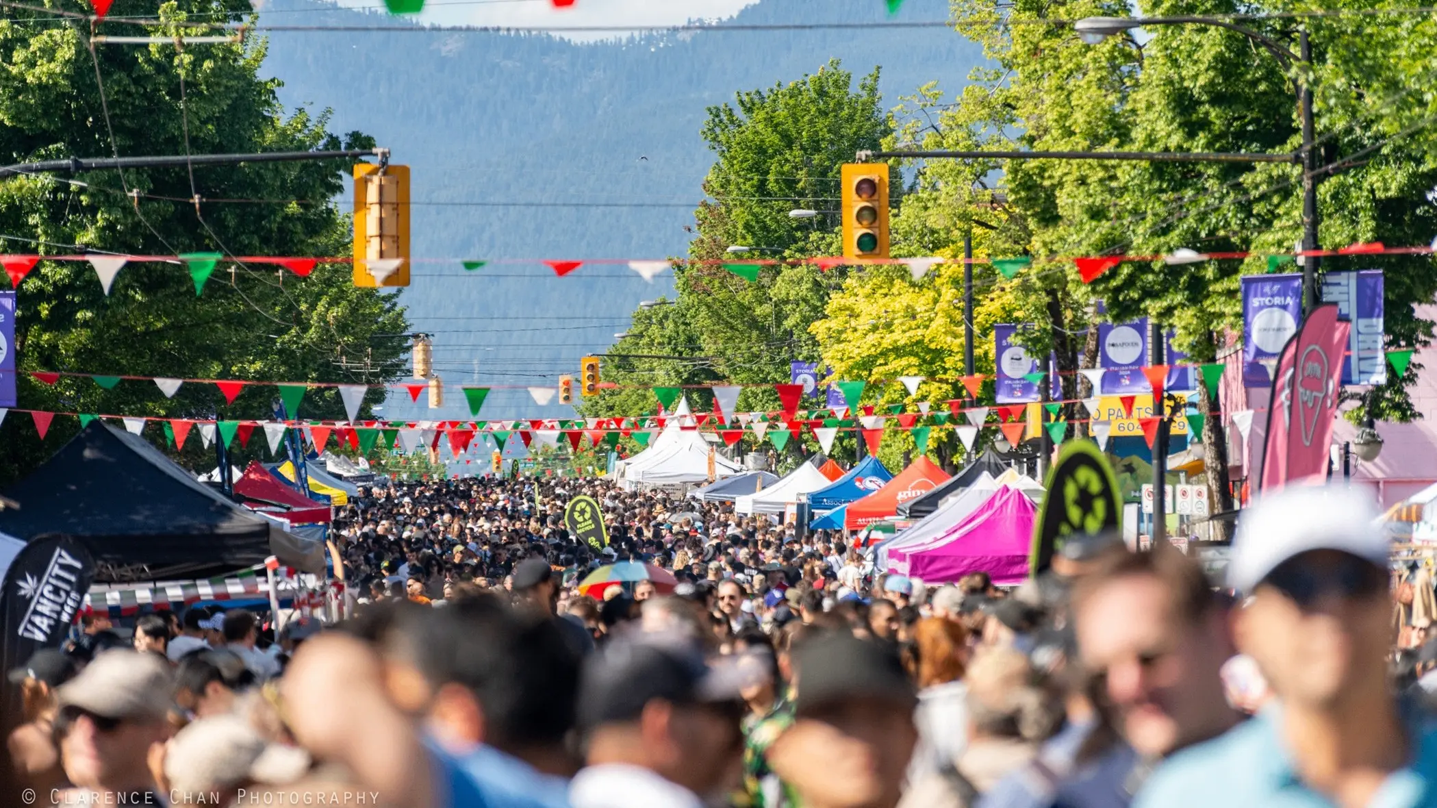 Crowded outdoor street market with colorful tents, green trees, and red, white, and green triangular flags overhead.