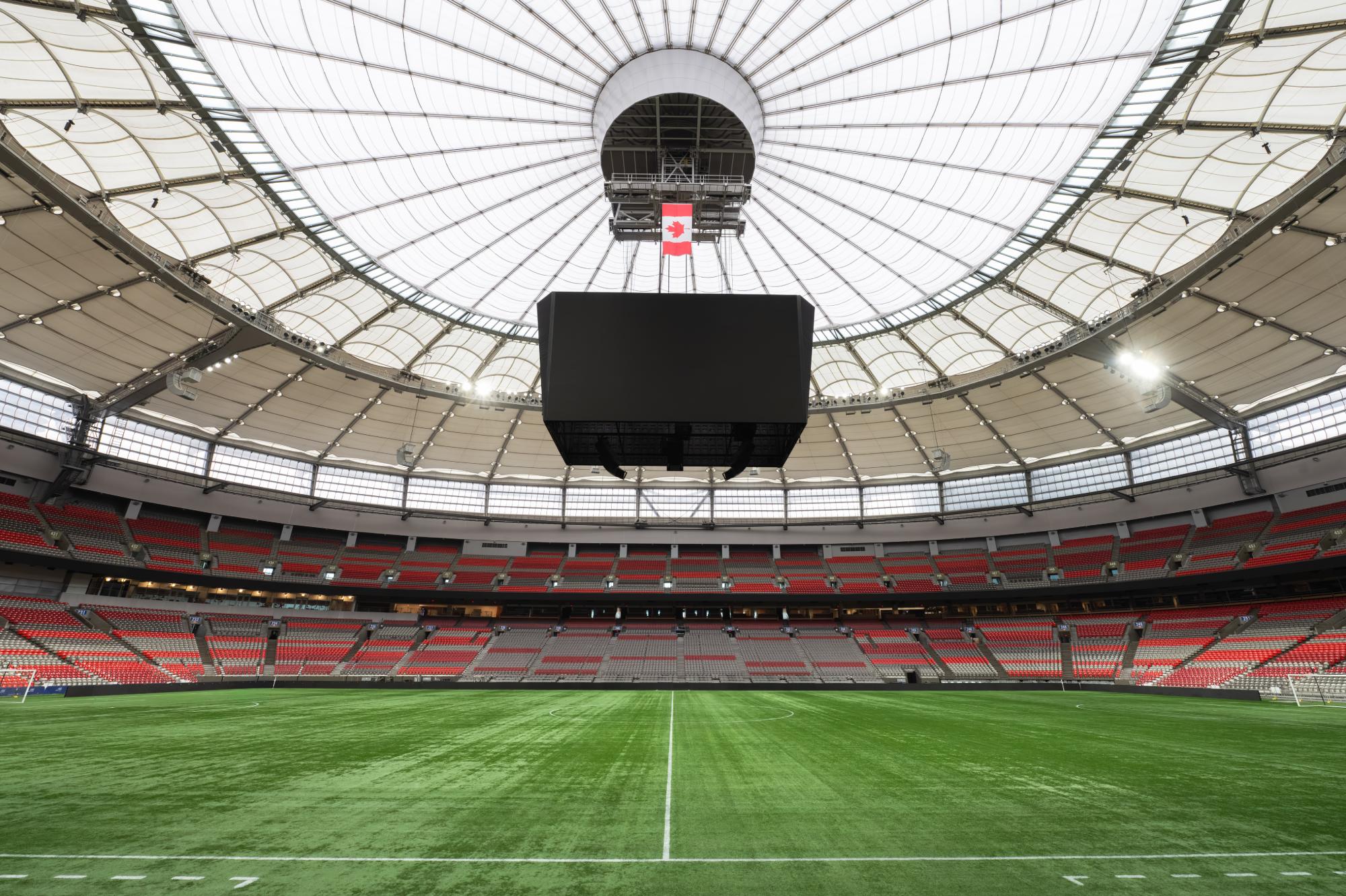 Empty indoor soccer stadium with green field, red and gray seats, and a large screen hanging from the white dome ceiling.