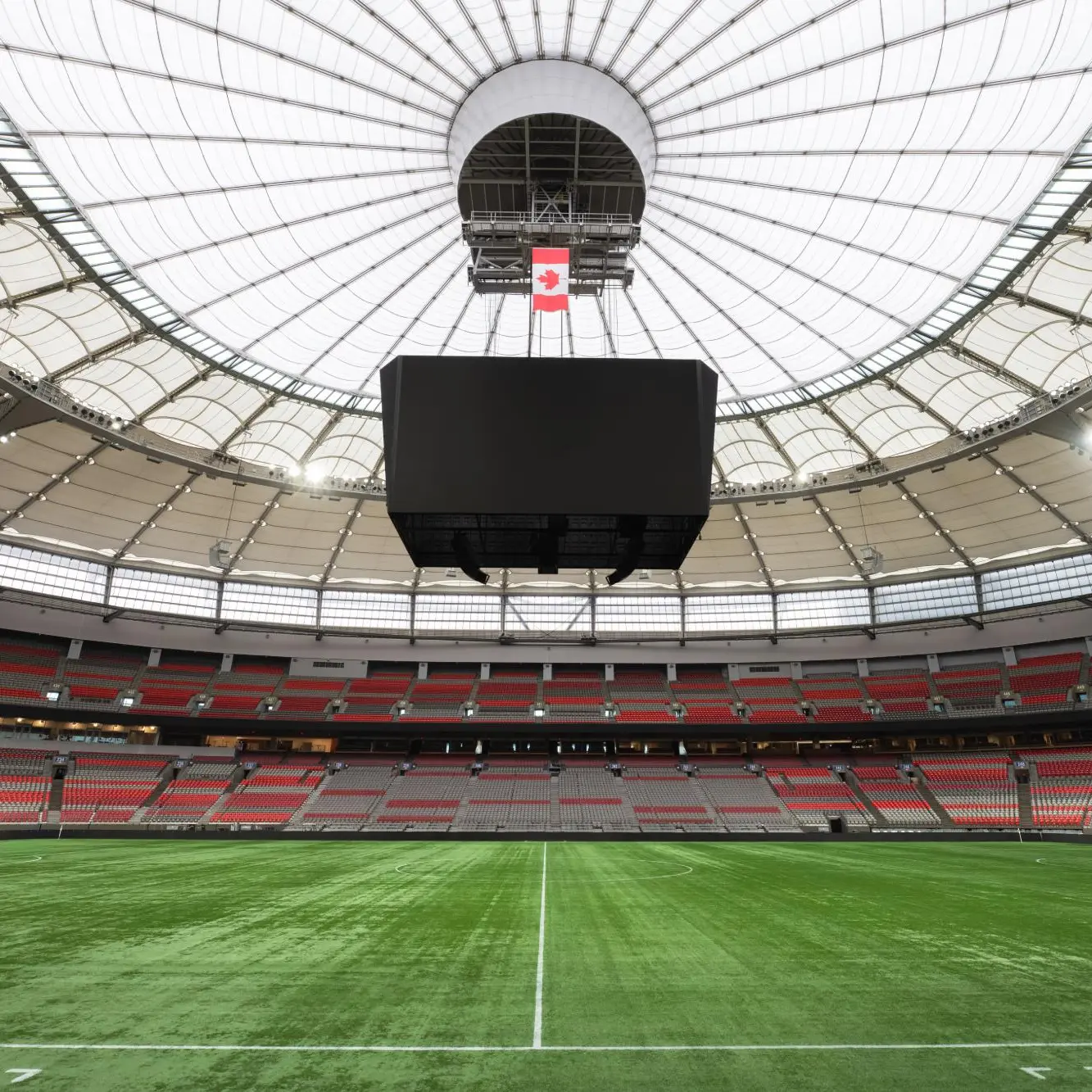 Empty indoor soccer stadium with green field, red and gray seats, and a large screen hanging from the white dome ceiling.