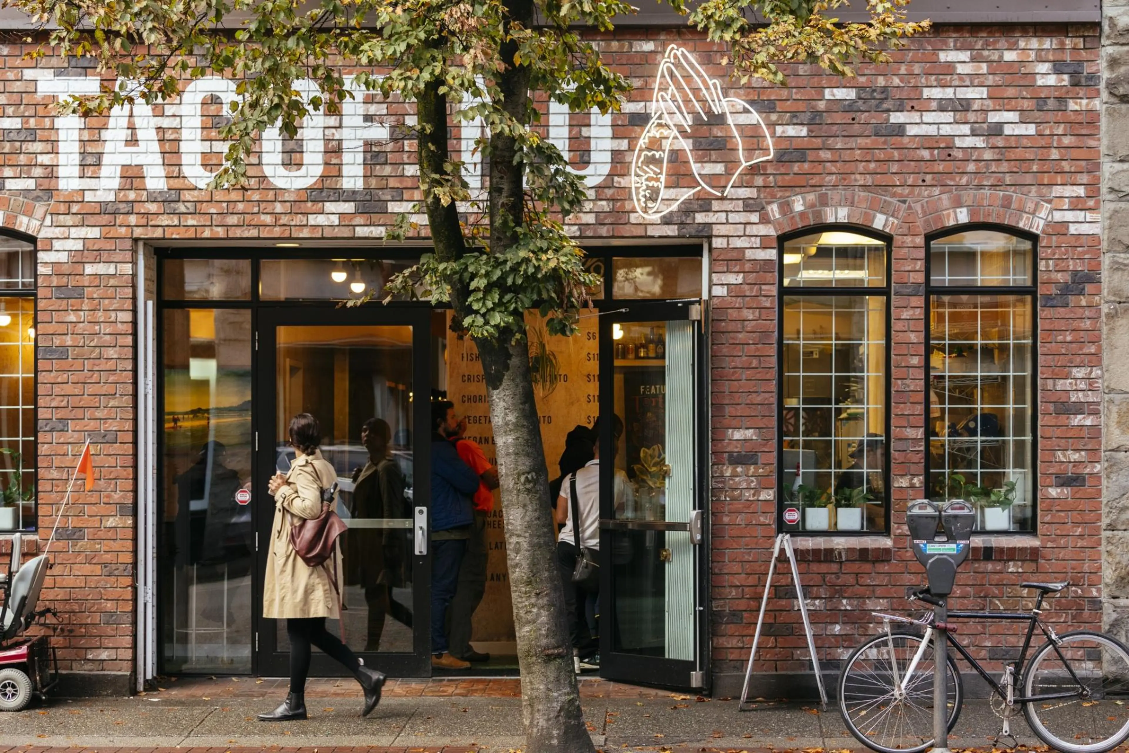 People entering a brick building with a sign showing praying hands holding a taco and partially visible text 'TACQUE'.