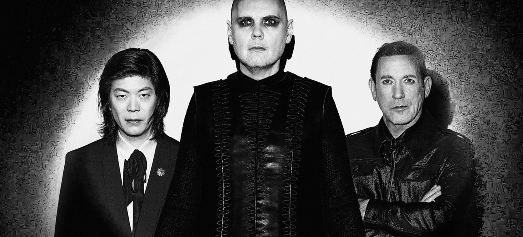 Three people standing against a wall with spotlight, wearing dark clothing, one with bald head and dark eye makeup.