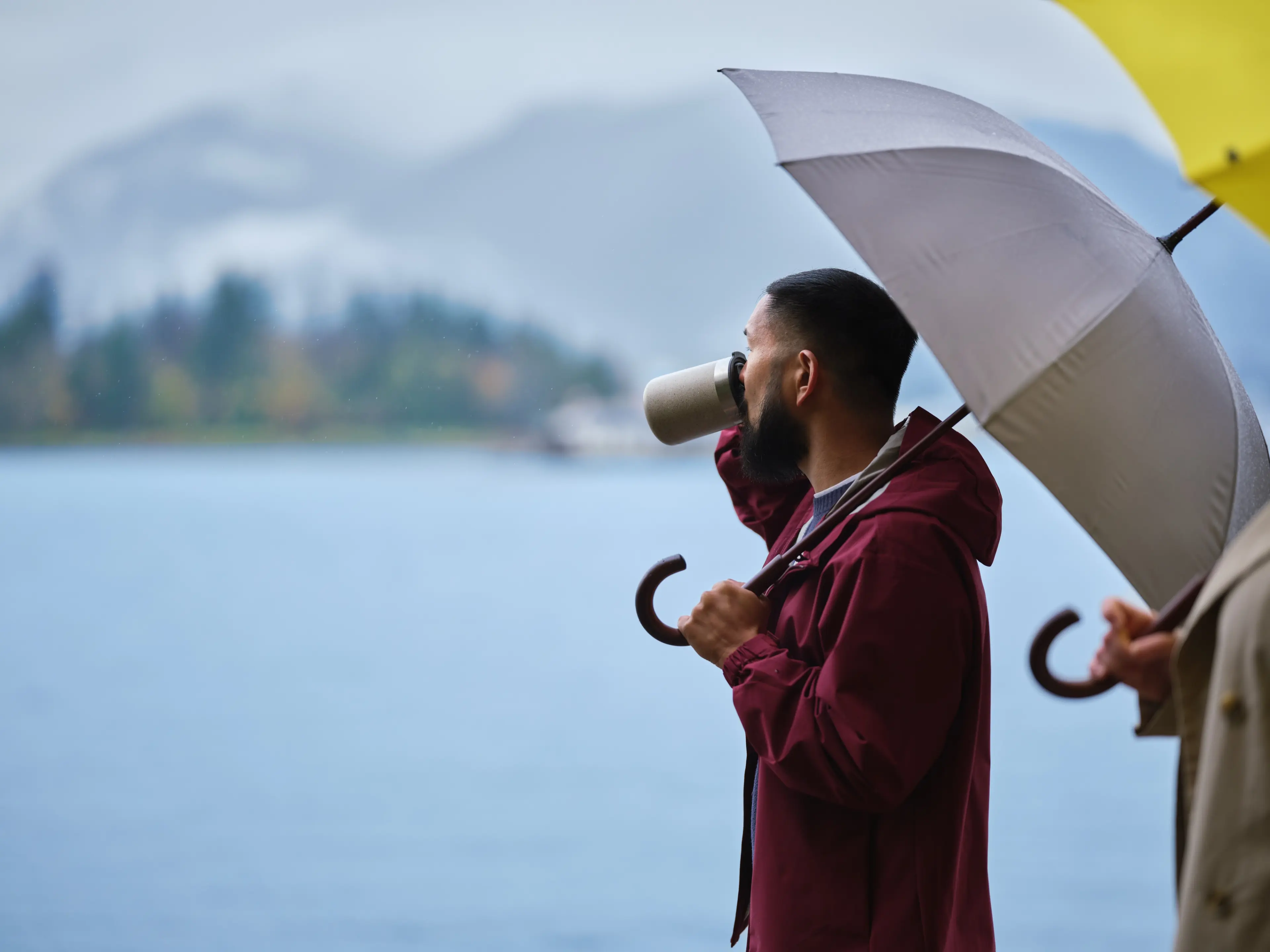 A person looking out of from Coal Harbour, drinking from a mug and holding an umbrella in Vancouver.