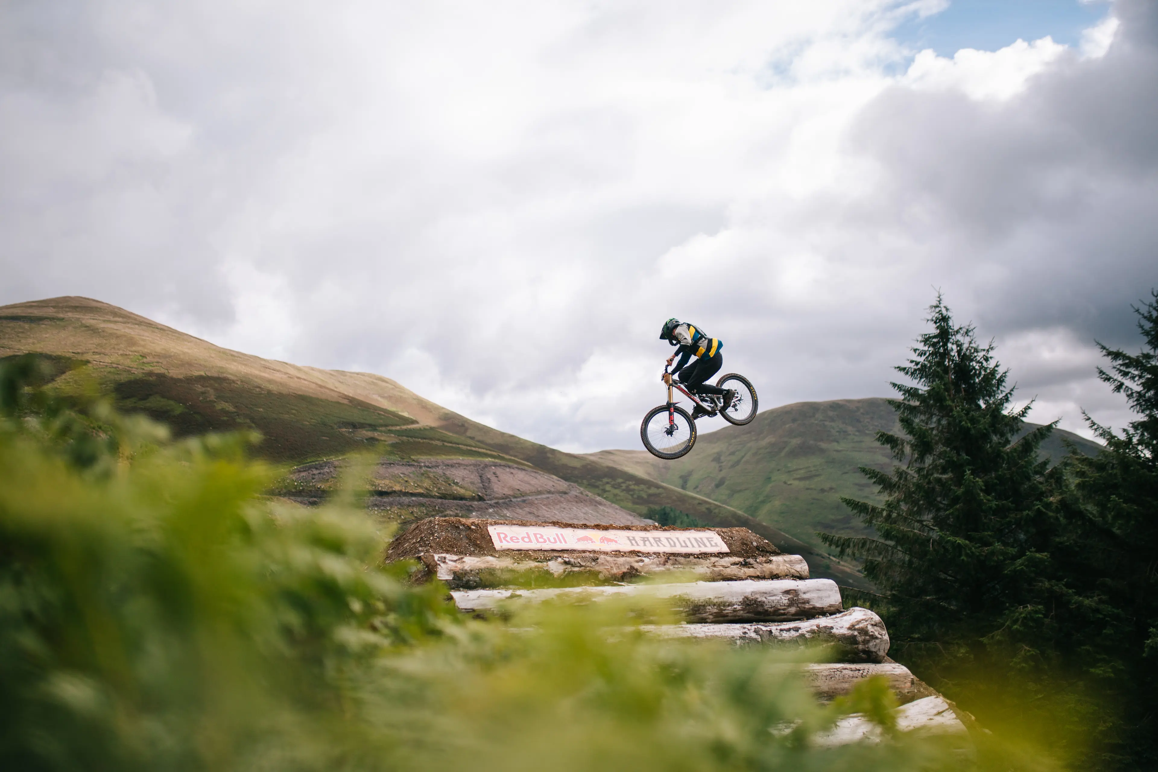 A person wearing a helmet jumps a mountain bike off a wooden ramp in a mountainous outdoor setting with cloudy sky.