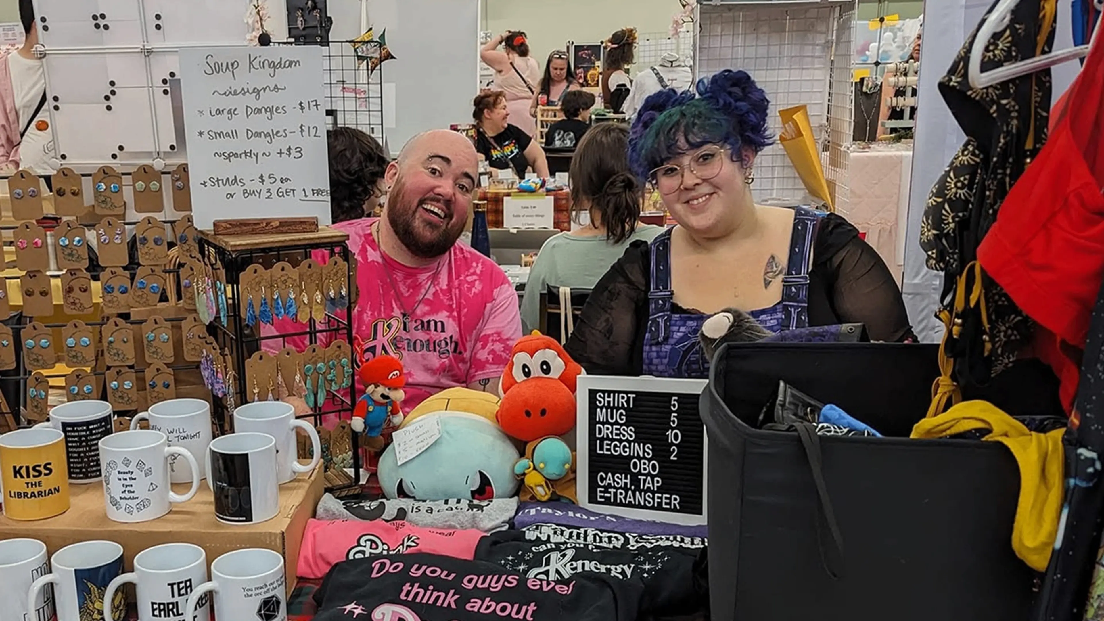 Two people sitting behind a table with mugs, earrings, plush toys, and clothing for sale at a market or fair.
