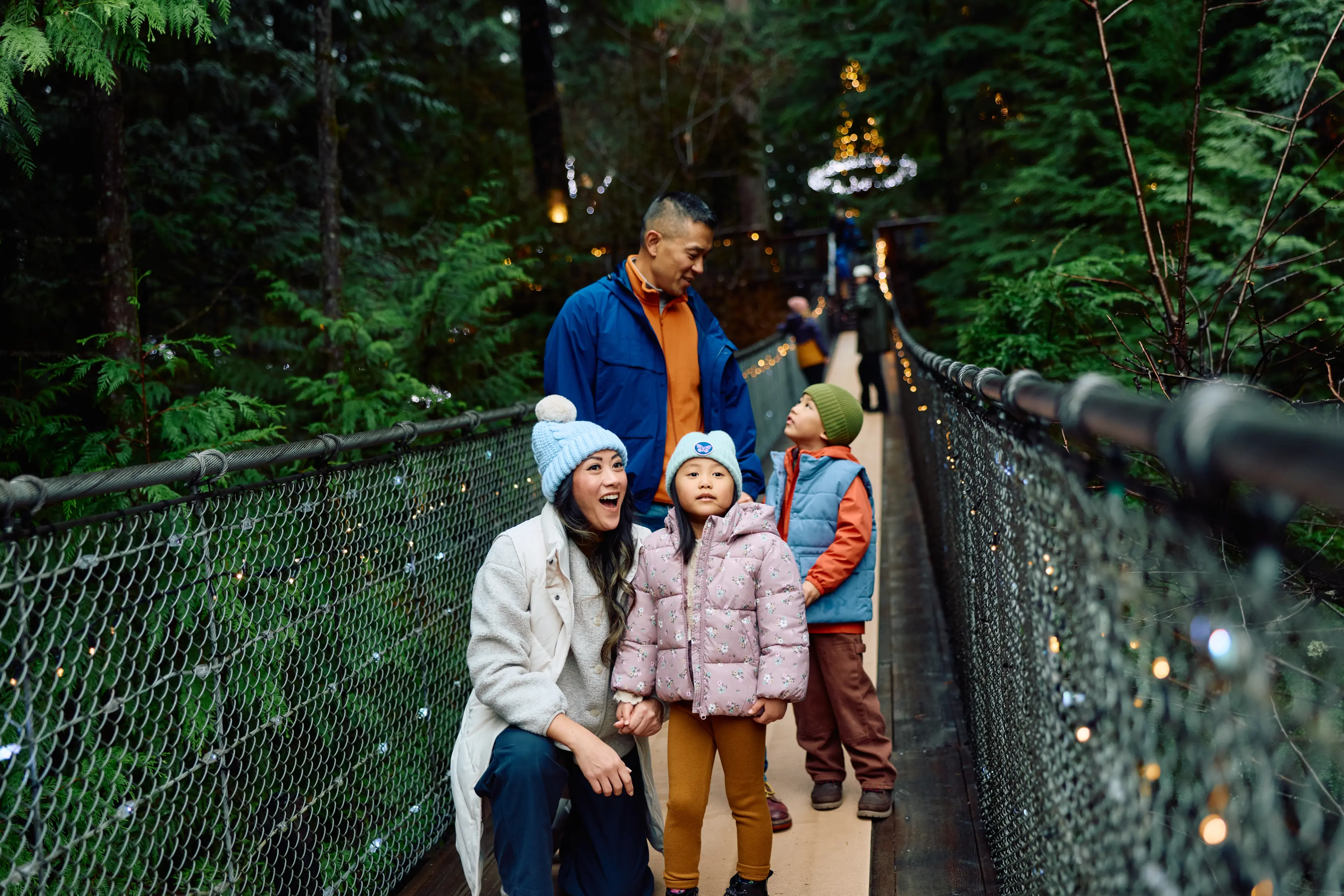 A family of four on a lit suspension bridge surrounded by green trees, with two children wearing winter jackets and hats.