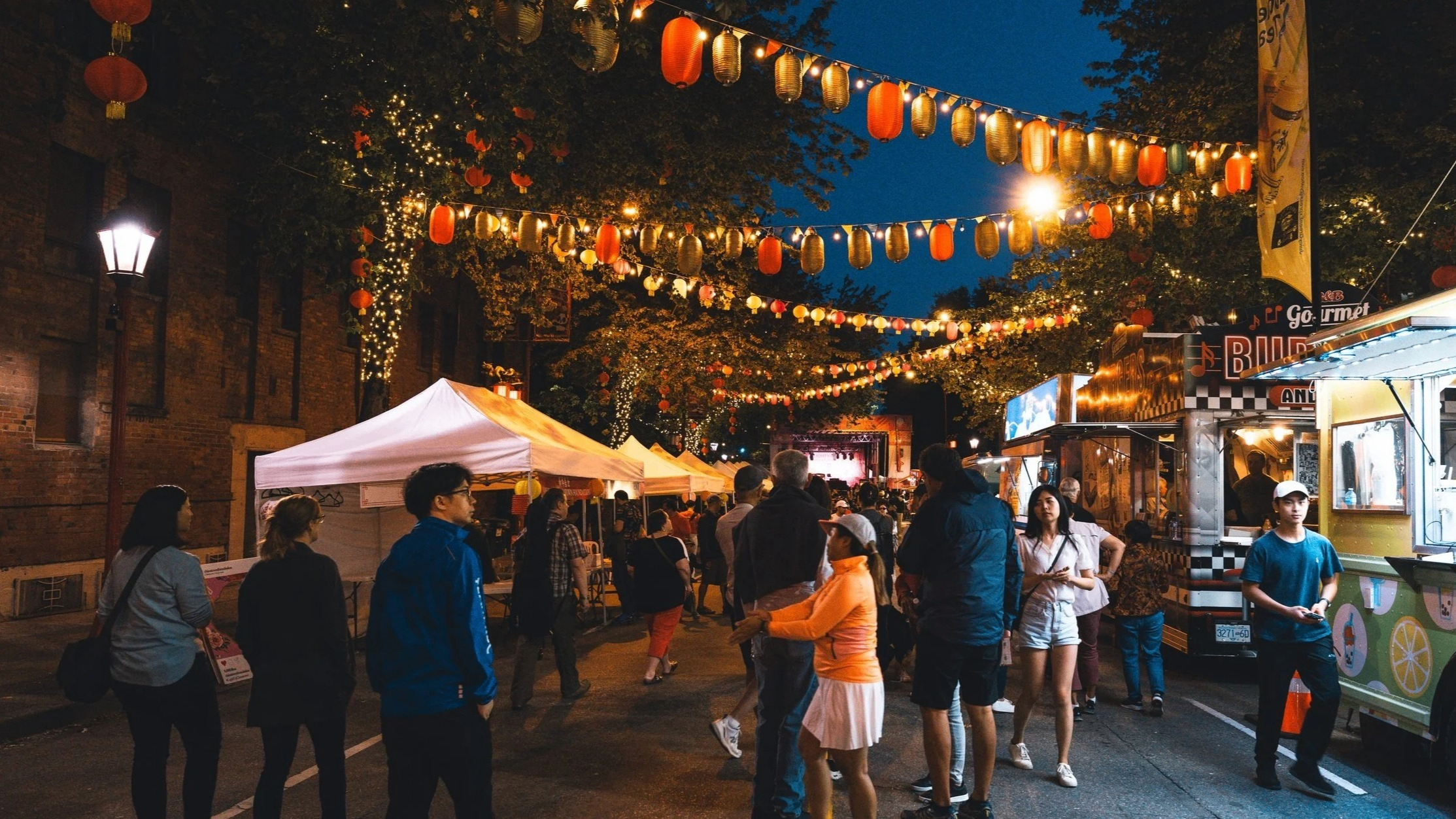 People walking and standing at a night street market decorated with hanging lanterns and string lights, with food trucks and tents on either side.