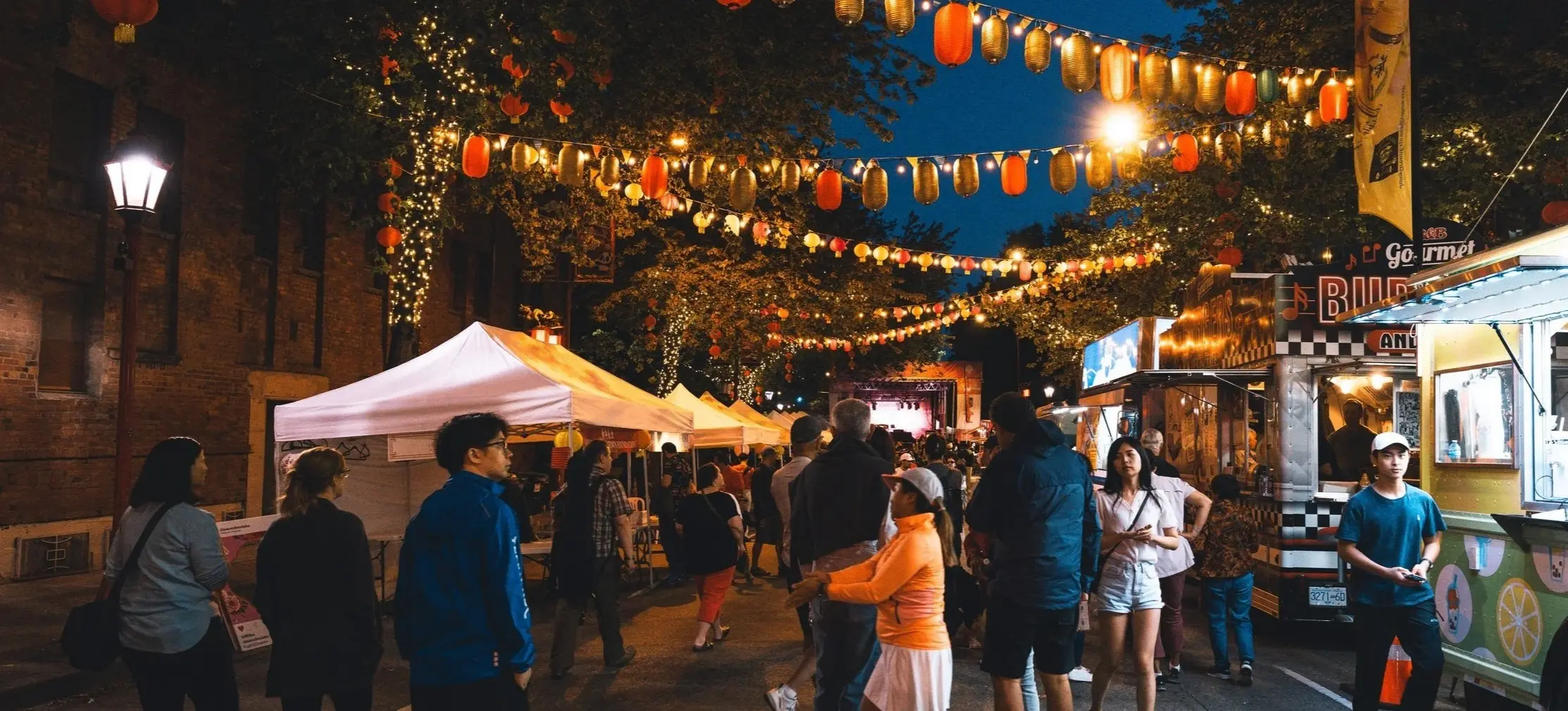 People walking and standing at a night street market decorated with hanging lanterns and string lights, with food trucks and tents on either side.