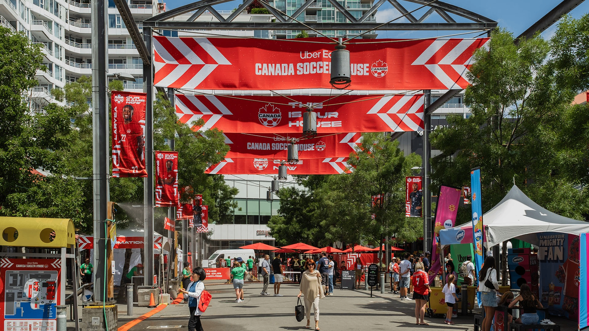 Outdoor event with red Canada Soccer House banners, people walking and tents on a sunny day in an urban area.