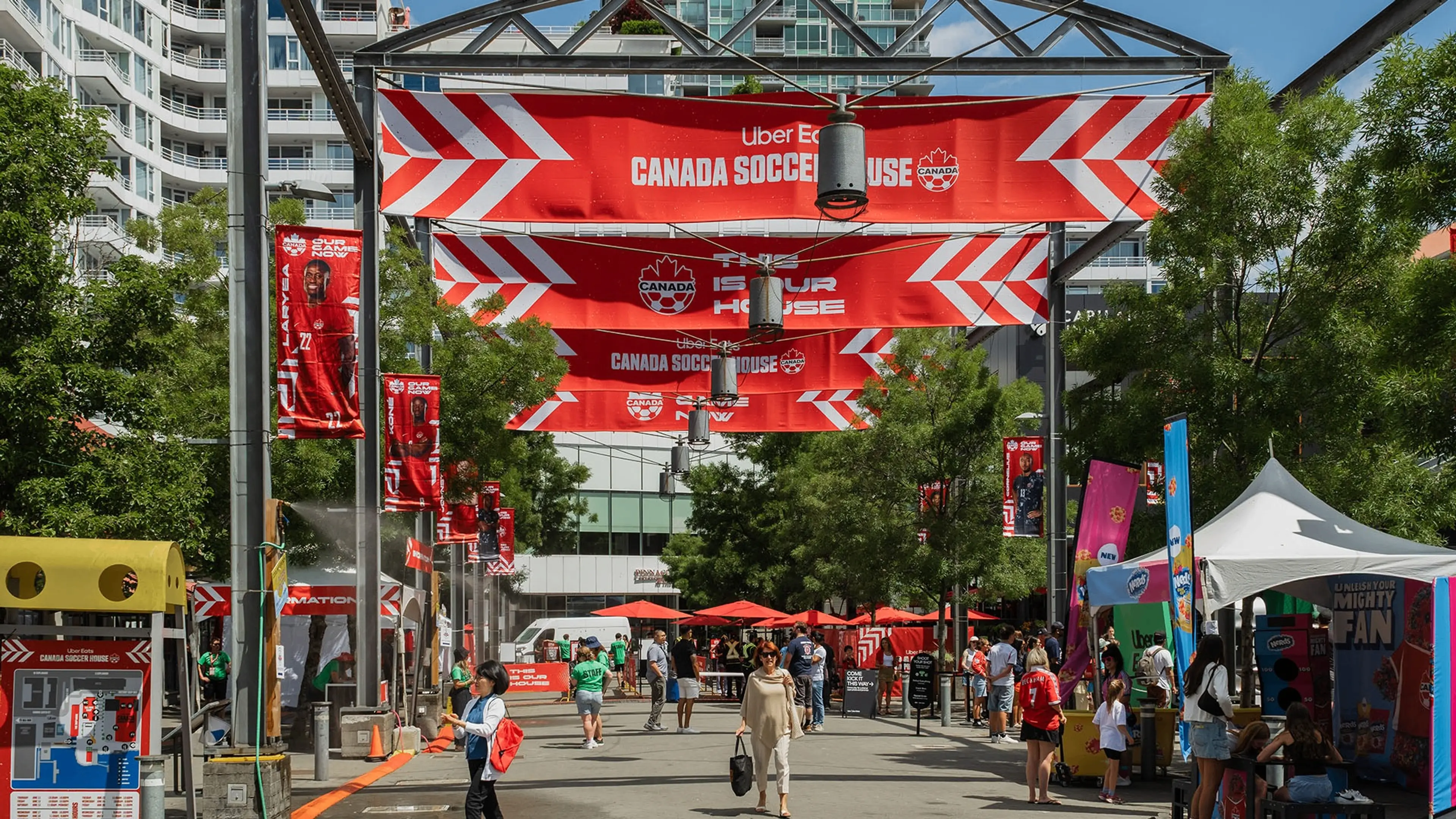 Outdoor event with red Canada Soccer House banners, people walking and tents on a sunny day in an urban area.