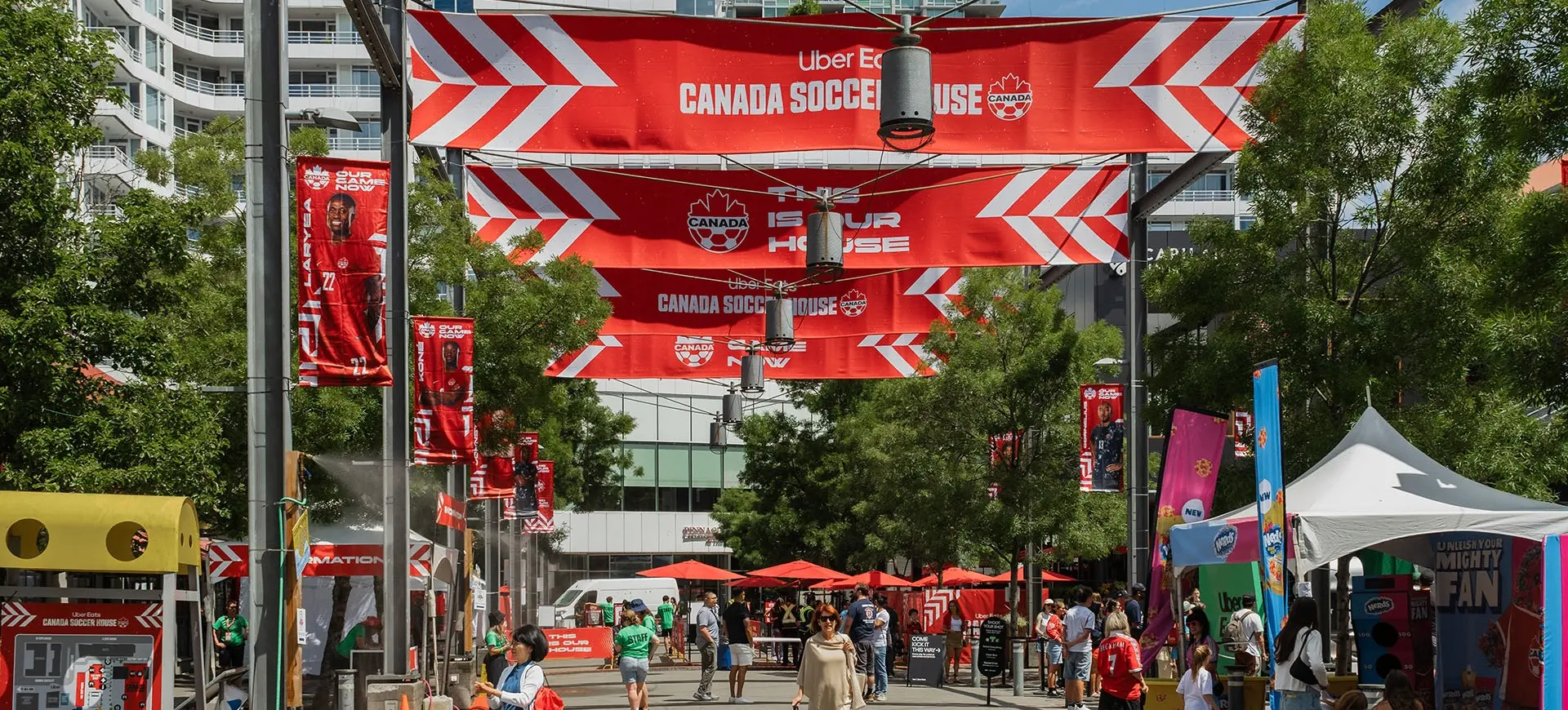 Outdoor event with red Canada Soccer House banners, people walking and tents on a sunny day in an urban area.