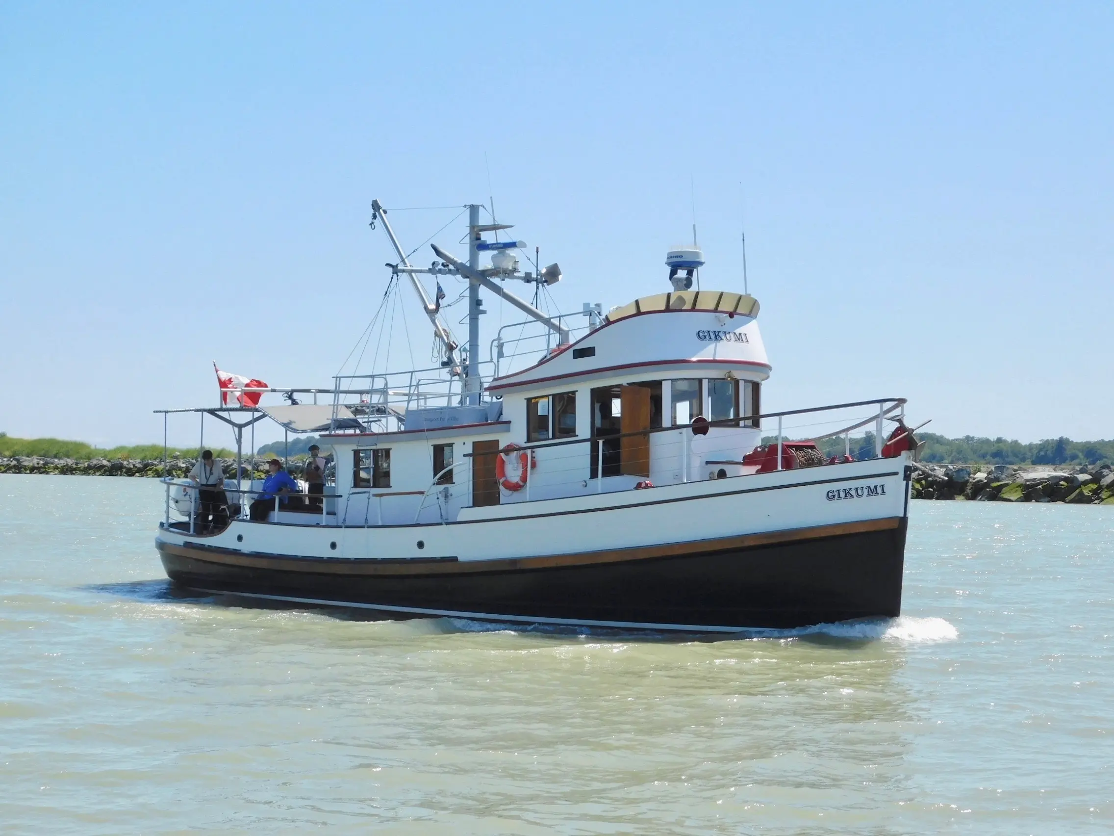 White and black boat named Gikumi with people on board sailing on calm water near rocky shore under clear blue sky.