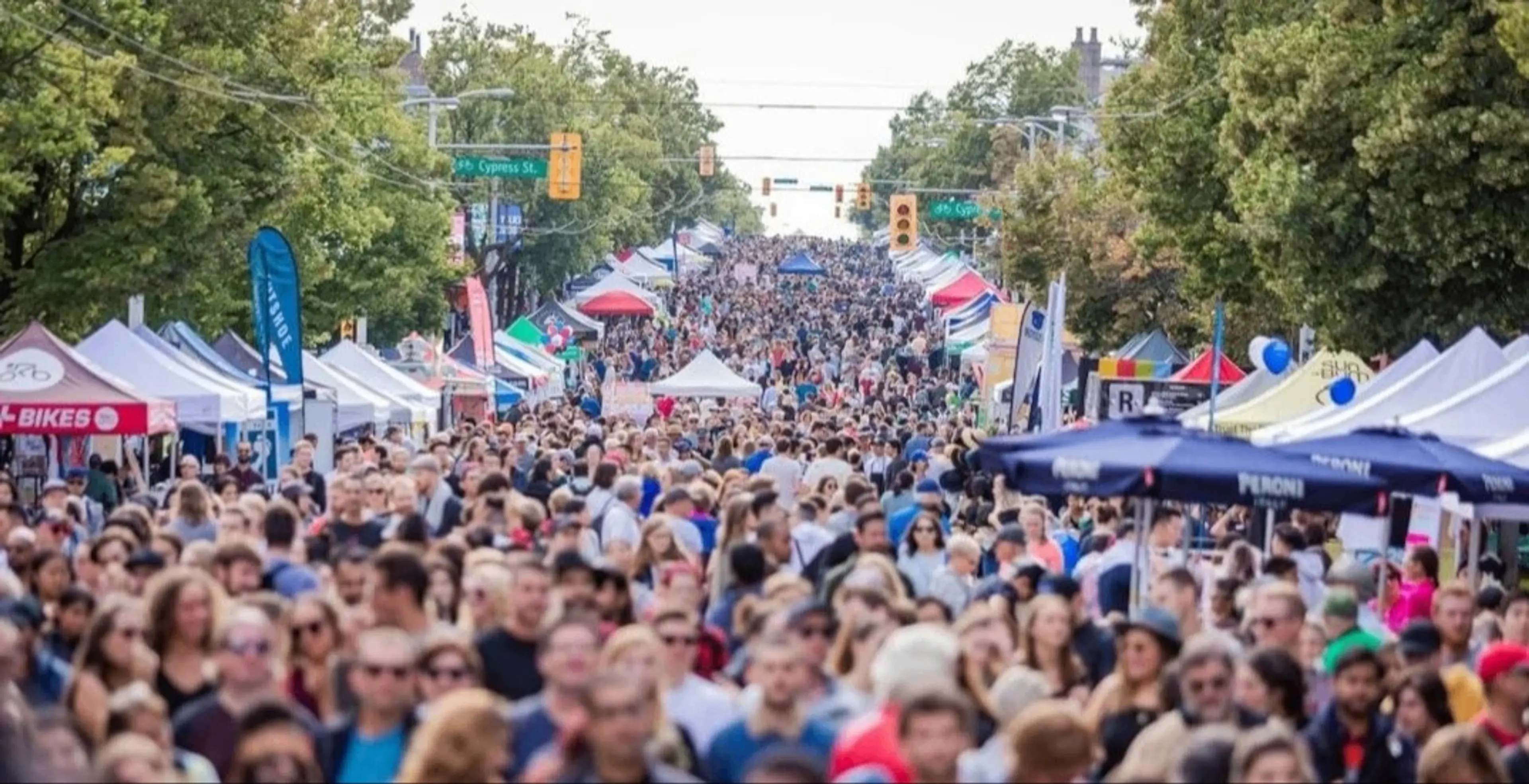 Crowded outdoor street market with many people walking between rows of vendor tents and trees lining the street.