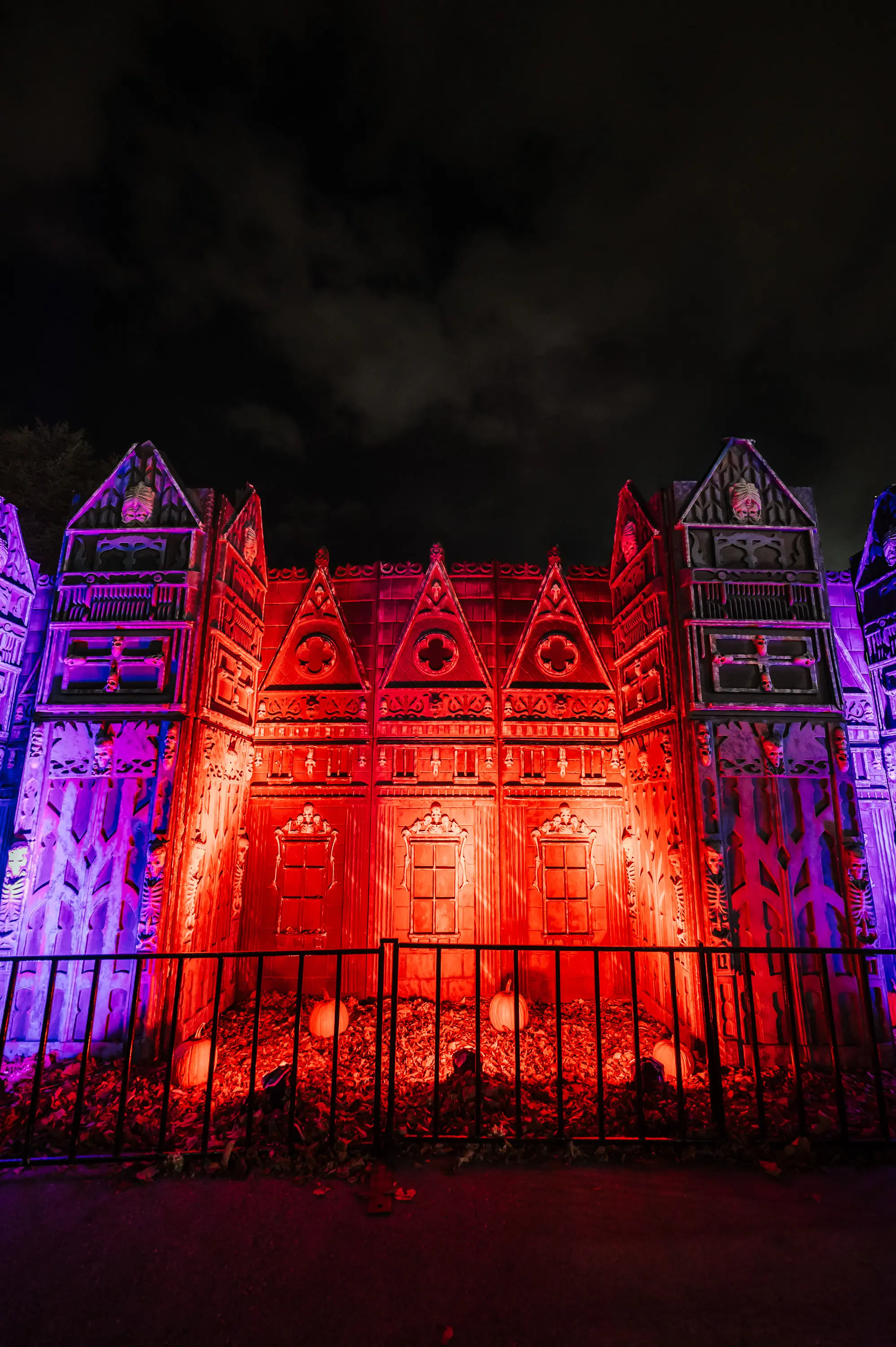 A Halloween-themed facade lit with red and purple lights, decorated with skeletons and pumpkins behind a black metal fence at night.