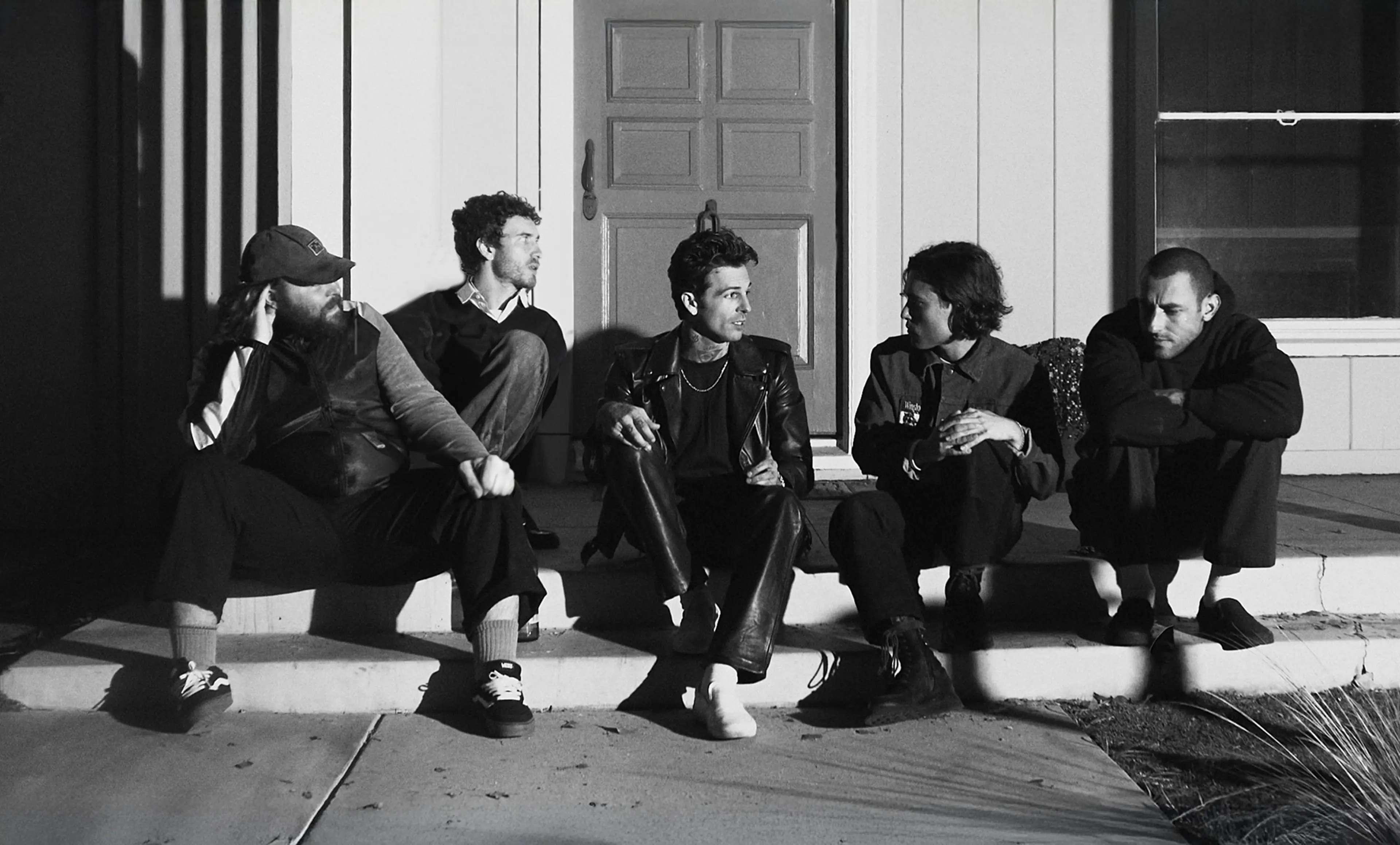 Five people sitting on outdoor steps in front of a door, engaged in conversation, in black and white.