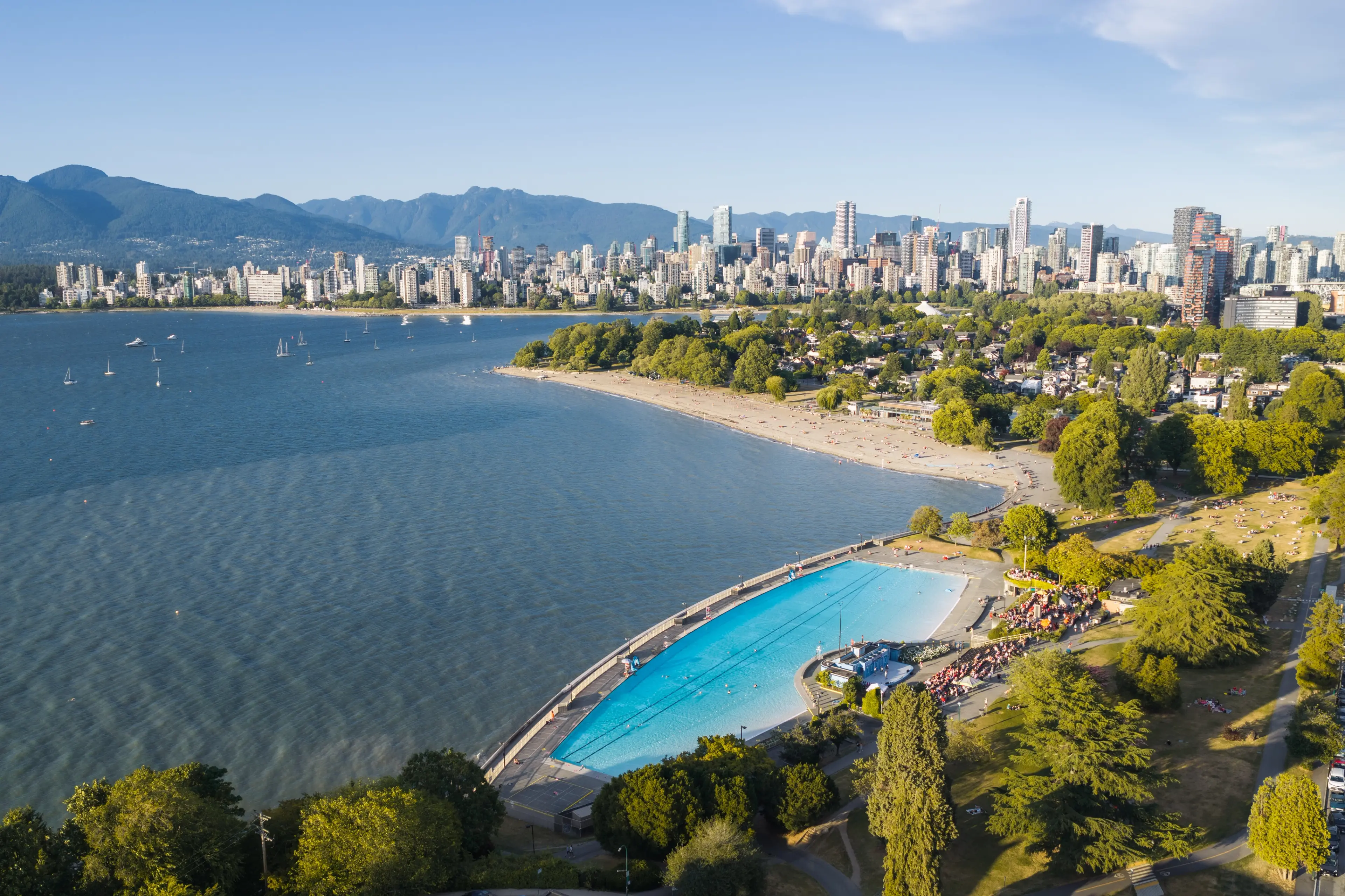 A large outdoor swimming pool near a beach with a city skyline and mountains in the background under a clear sky.