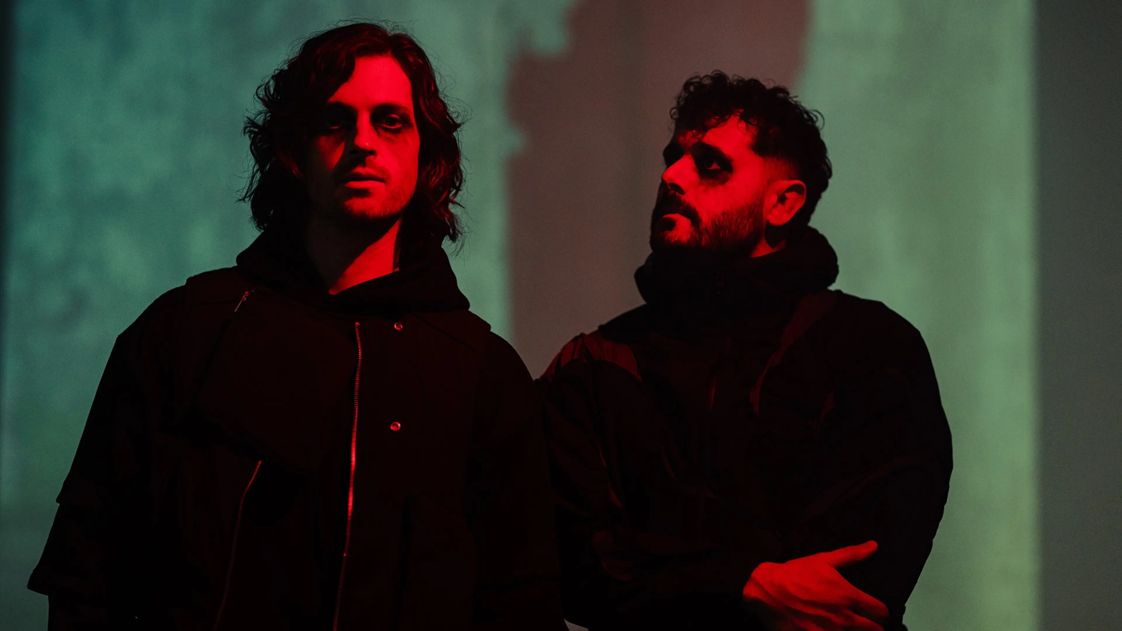 Two people with dark makeup and black clothing, lit by red light against a greenish background.