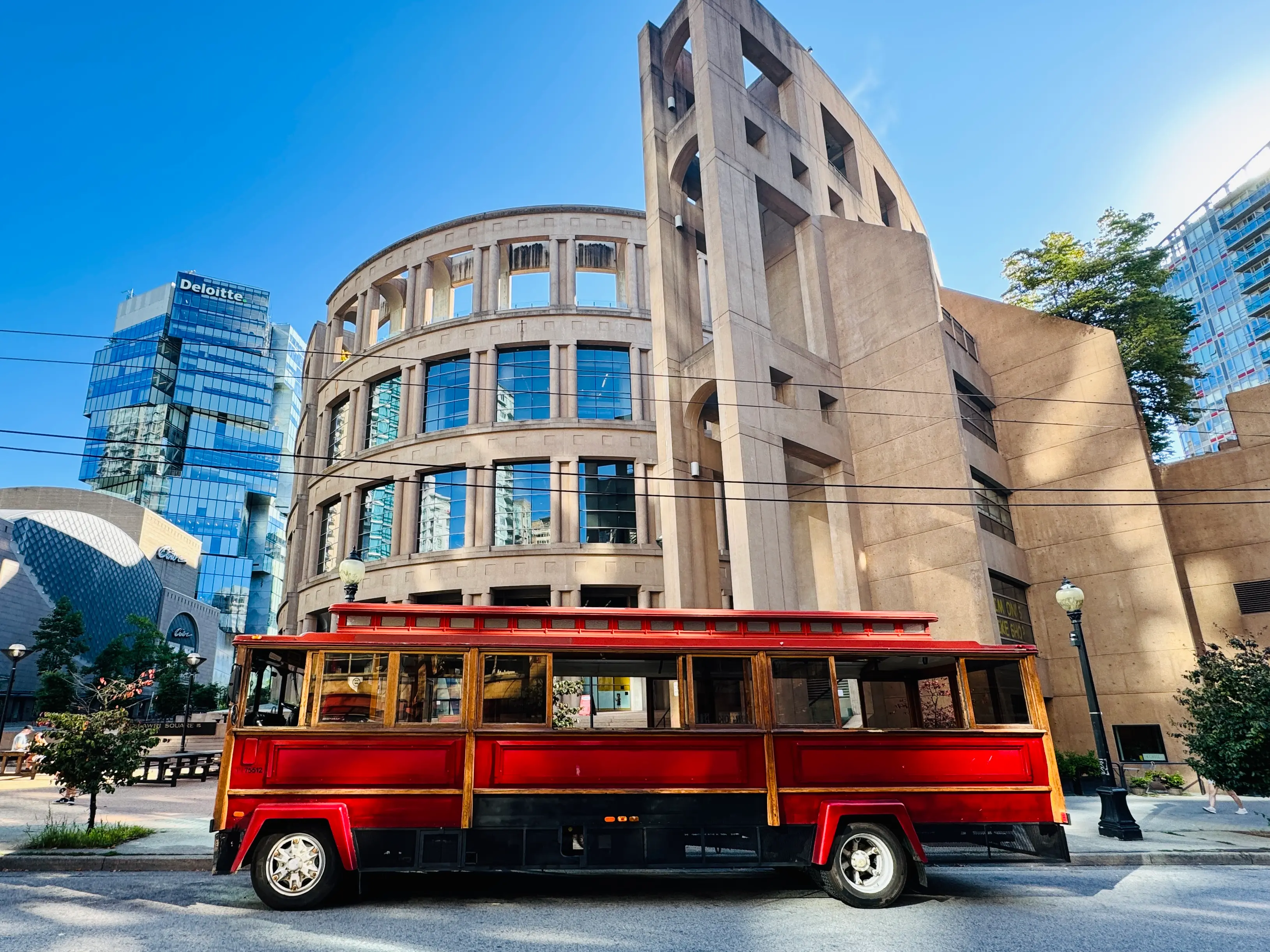 Red trolley bus parked on a city street with the Vancouver Library and glass skyscrapers in the background.