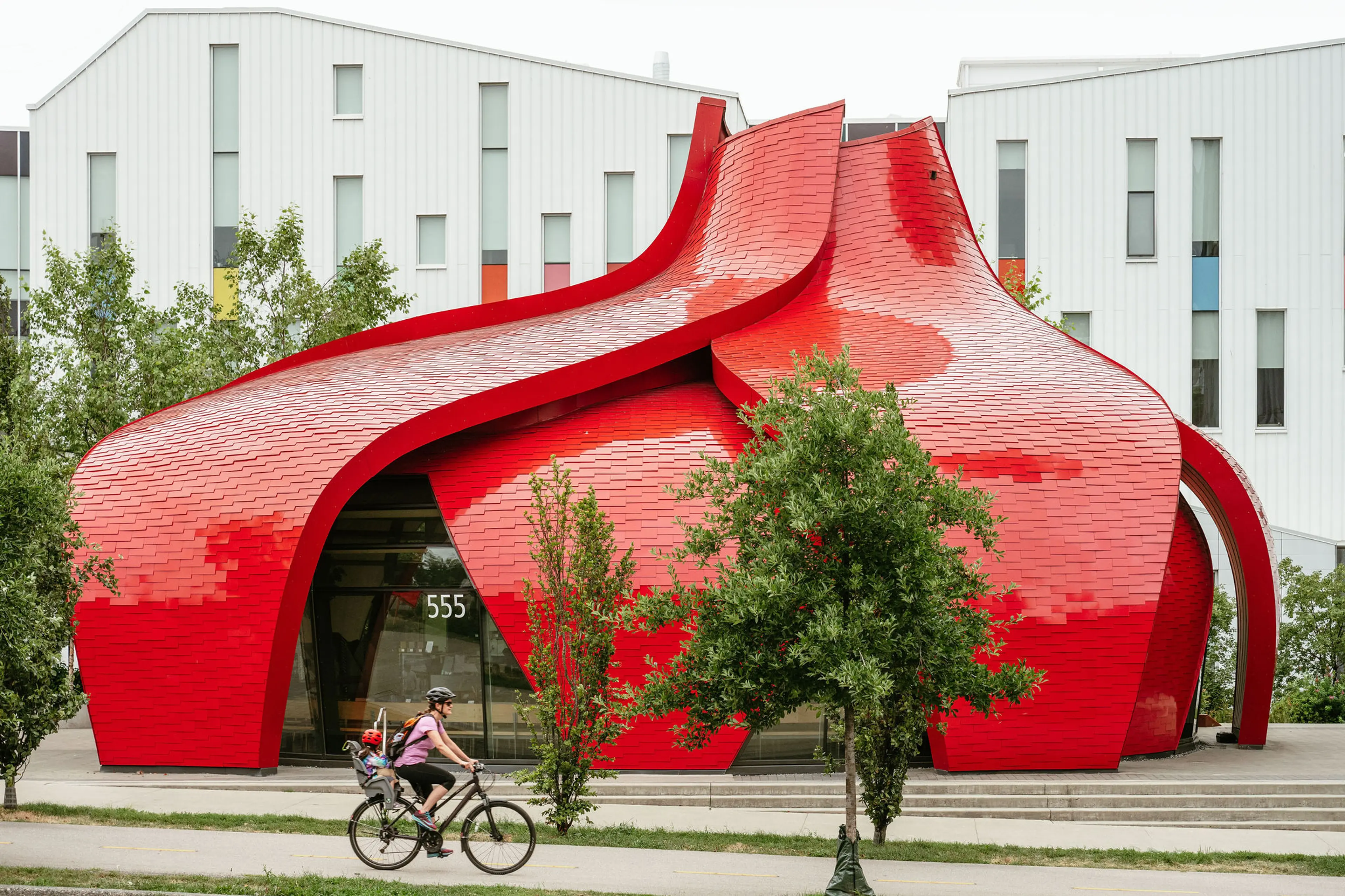 Person wearing a helmet rides a bicycle with a child seat past a modern red building with curved, flowing shapes and a tiled surface.