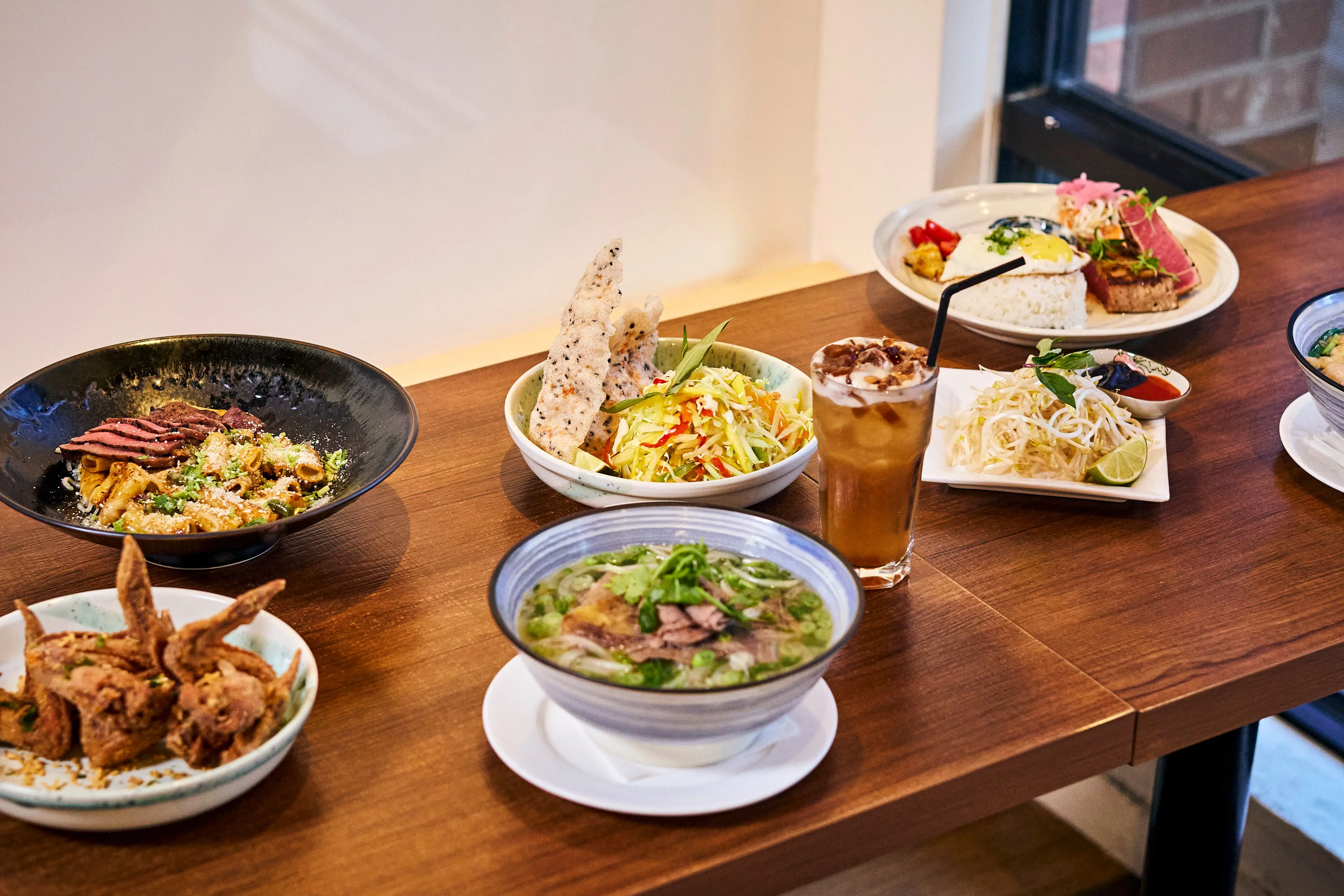 Table with various Asian dishes including noodle soup, fried chicken wings, salad with rice crackers, iced drink, and rice with meat and egg at the restaurant The Saigonville in Vancouver.