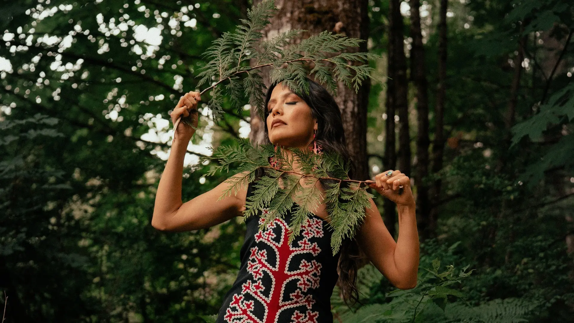 Person with long dark hair holding green fern branches in front of face, standing in a forest with eyes closed.