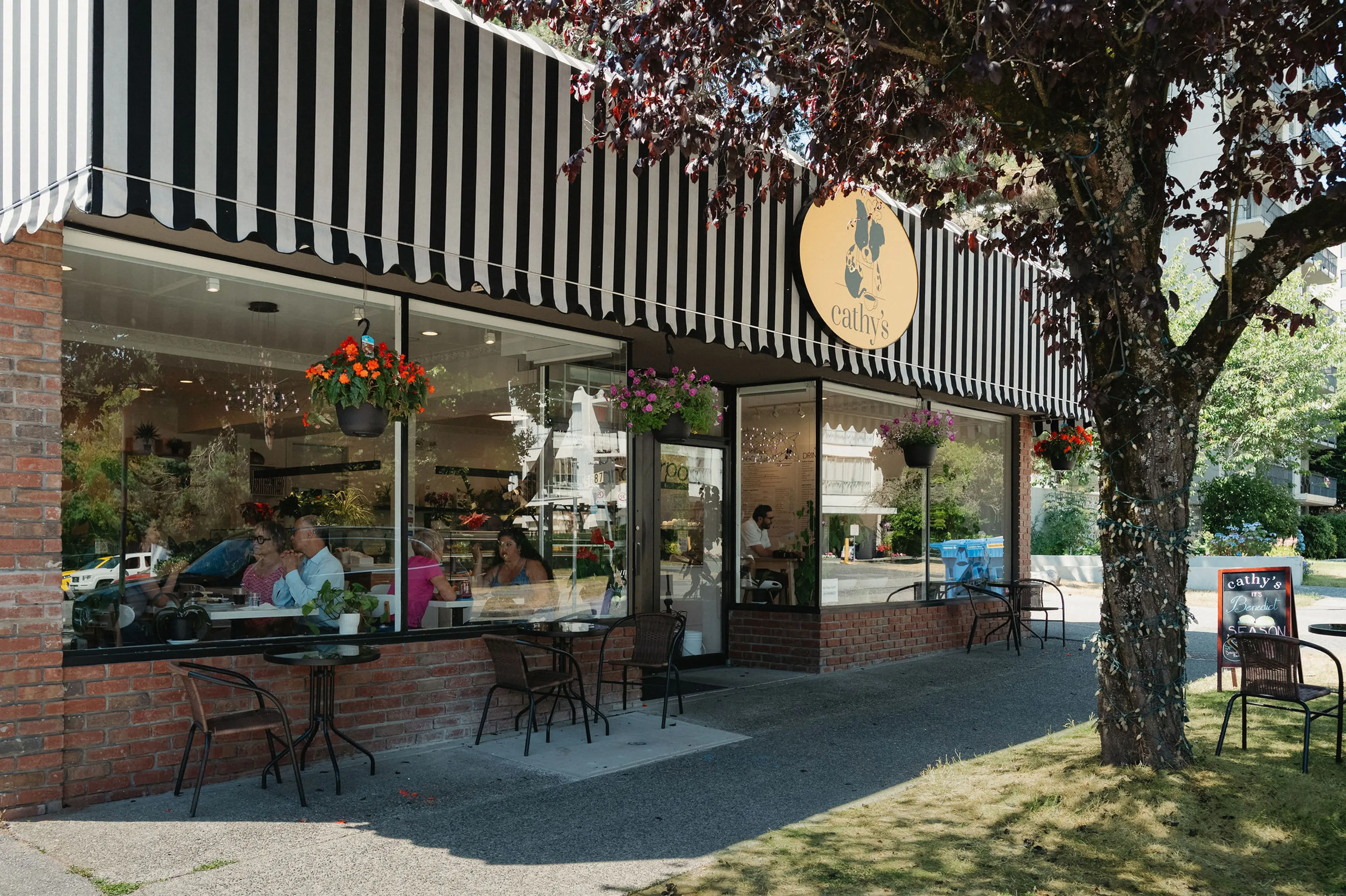 Cafe with black and white striped awning, hanging flower pots, outdoor tables and chairs, and people sitting inside.