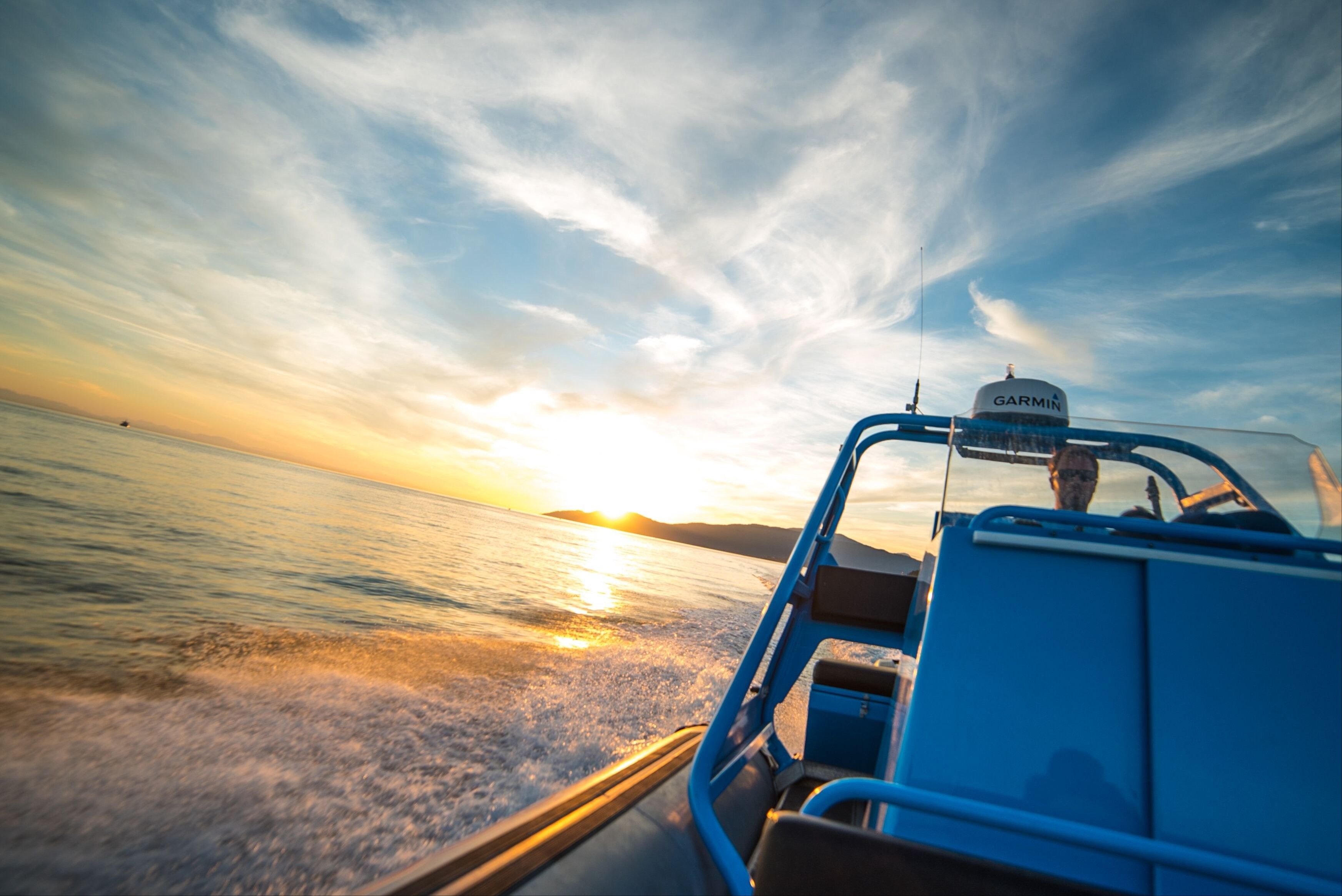 Person driving a blue motorboat on calm water during sunset with partly cloudy sky.