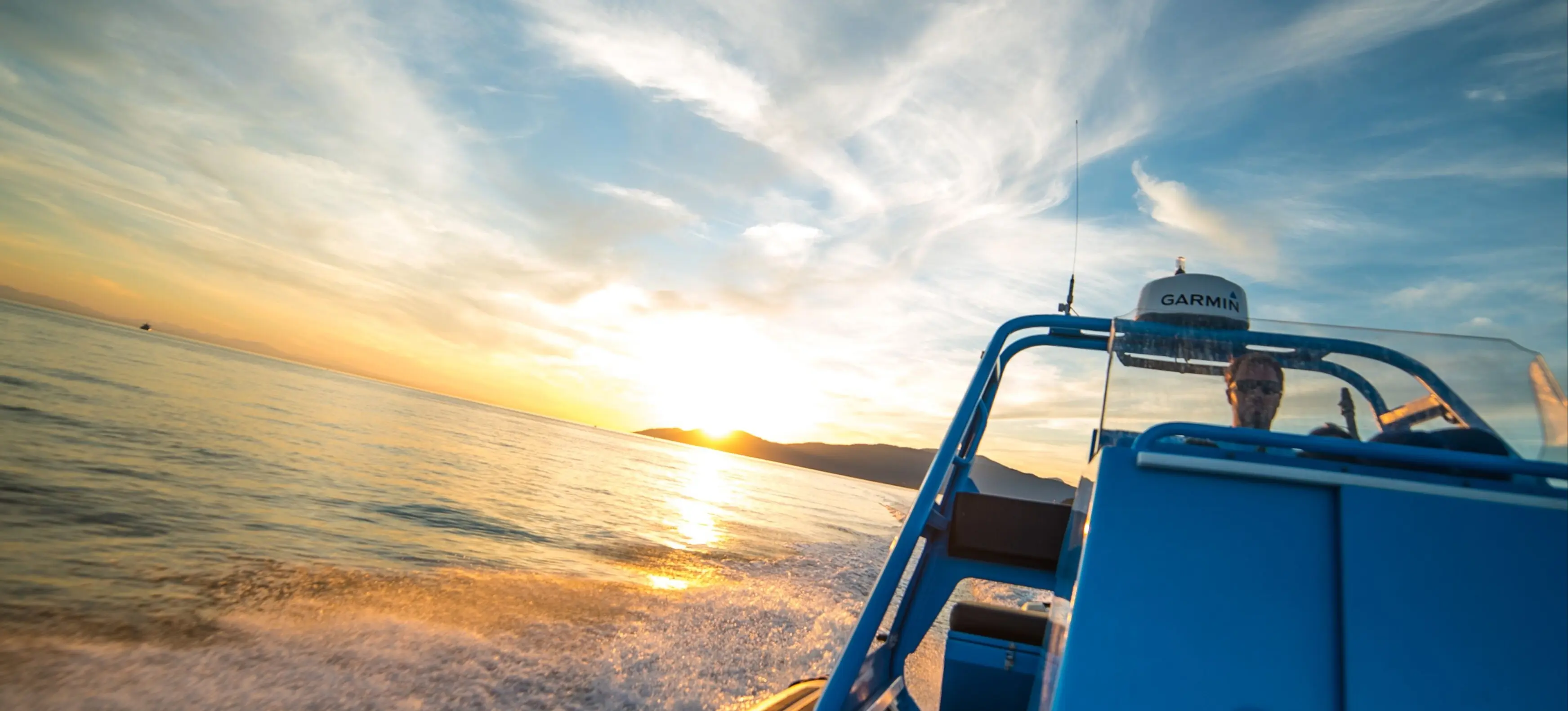 Person driving a blue motorboat on calm water during sunset with partly cloudy sky.