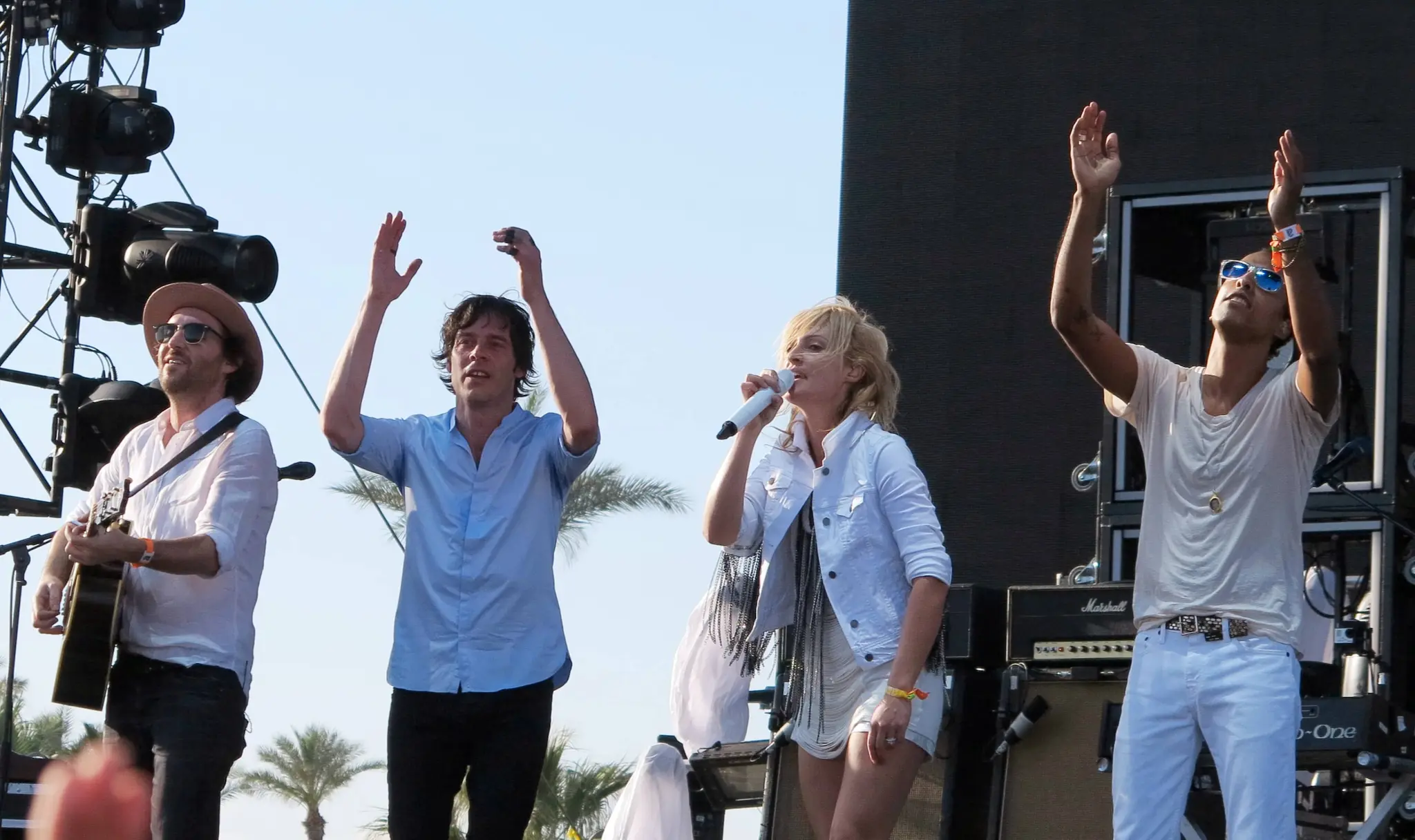 Four musicians performing on an outdoor stage, one playing guitar, one singing into a microphone, and two raising their hands.