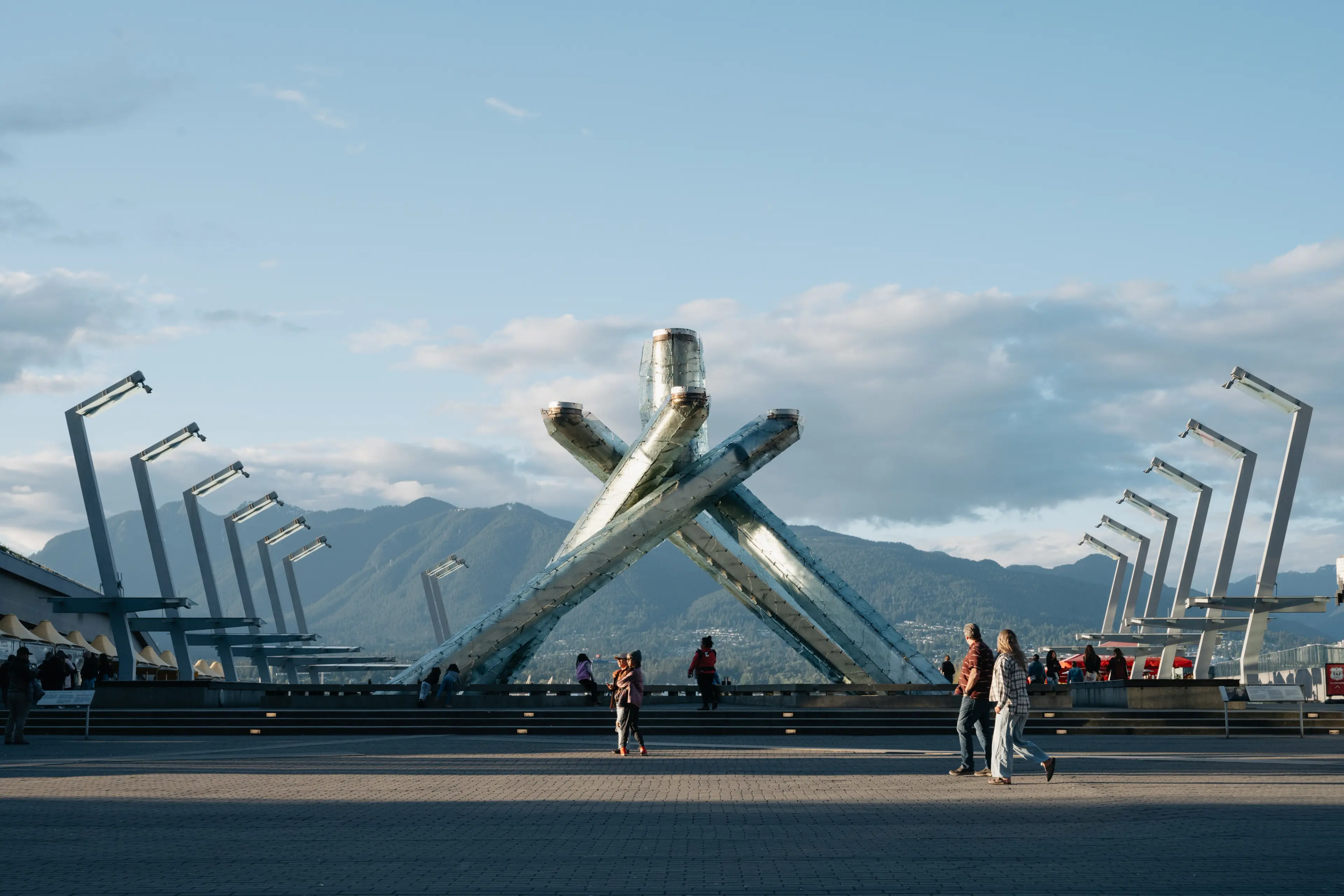 Large metallic Olympic cauldron sculpture with people walking nearby and mountains in the background under a blue sky.