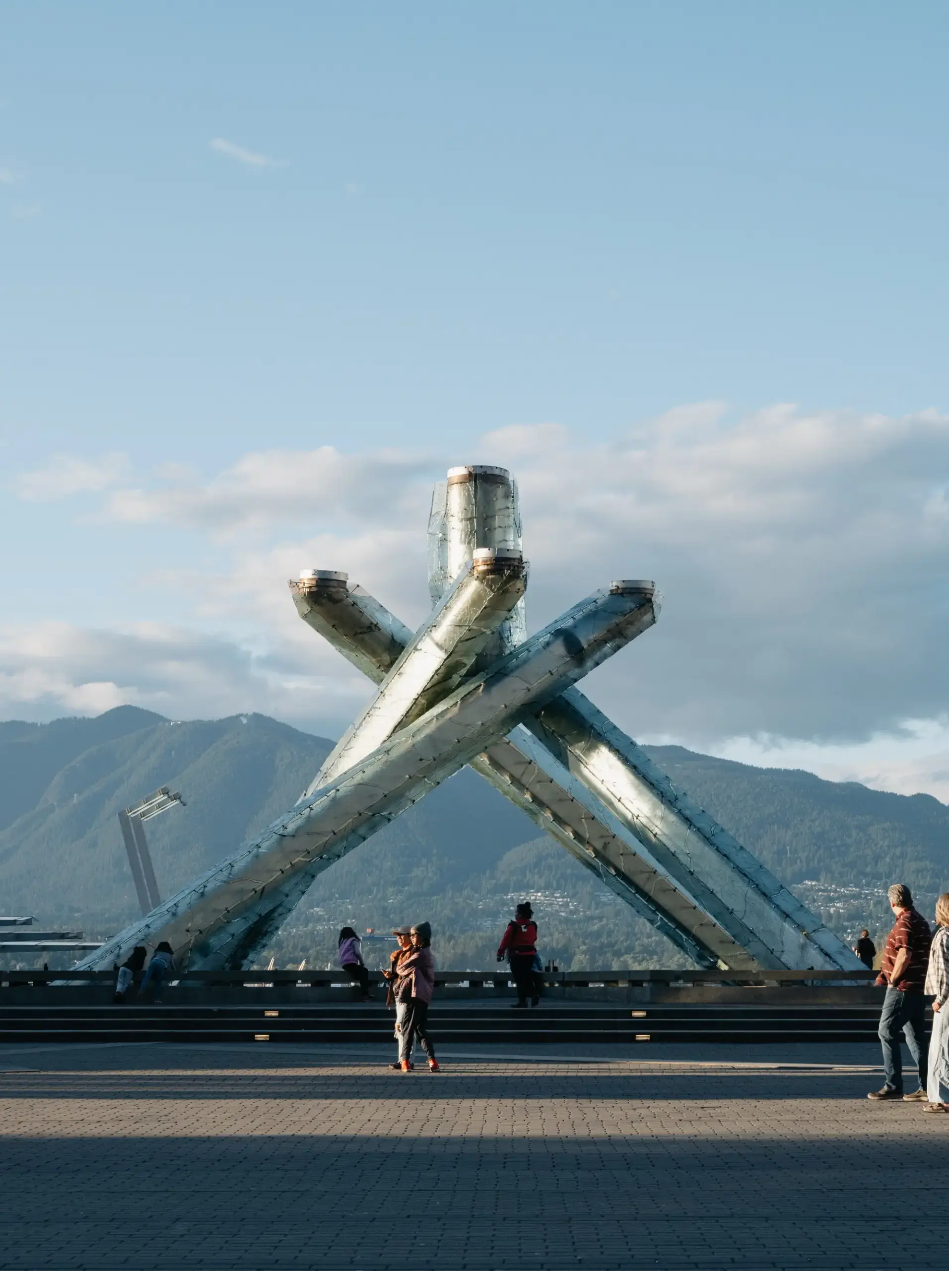 Large metallic Olympic cauldron sculpture with people walking nearby and mountains in the background under a blue sky.