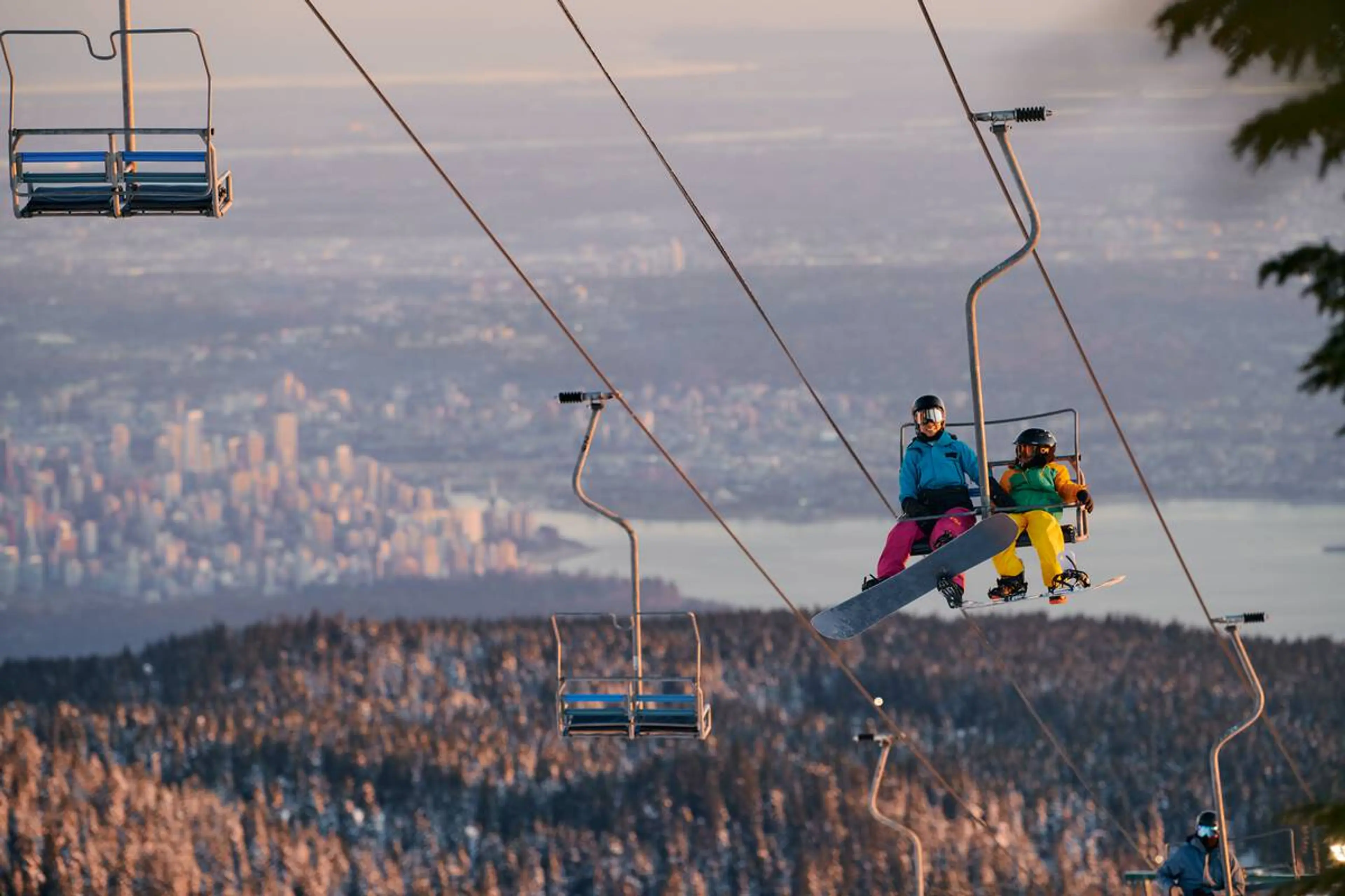 Two snowboarders ride a chairlift at Cypress Mountain with the city of Vancouver in the background.