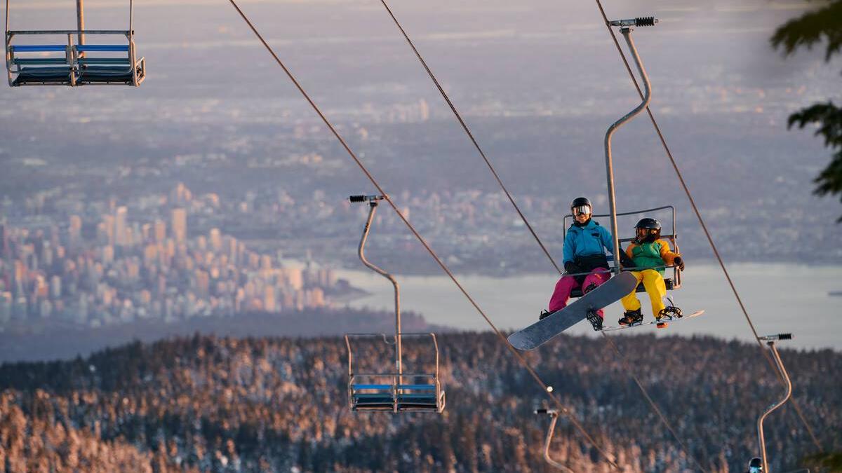 Two snowboarders ride a chairlift at Cypress Mountain with the city of Vancouver in the background.