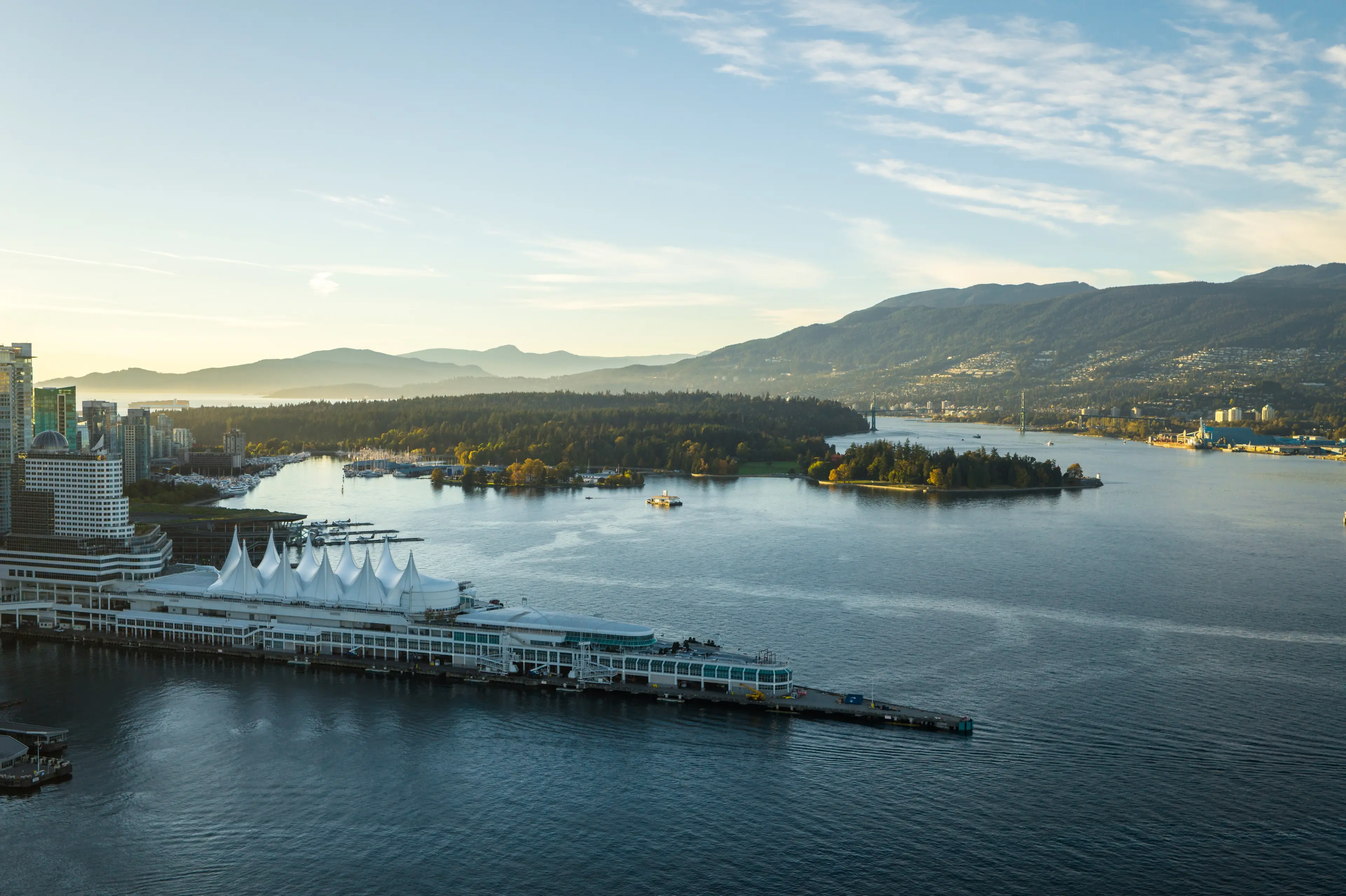 Waterfront cityscape with a pier featuring white tent-like structures, forested islands, and mountains in the background under a partly cloudy sky.