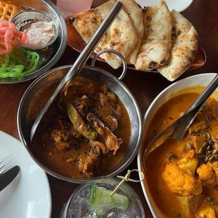 Table with Indian dishes including curry with green chili, cauliflower curry, naan bread, colorful jalebi sweets, and a glass of cucumber water.