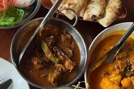 Table with Indian dishes including curry with green chili, cauliflower curry, naan bread, colorful jalebi sweets, and a glass of cucumber water.