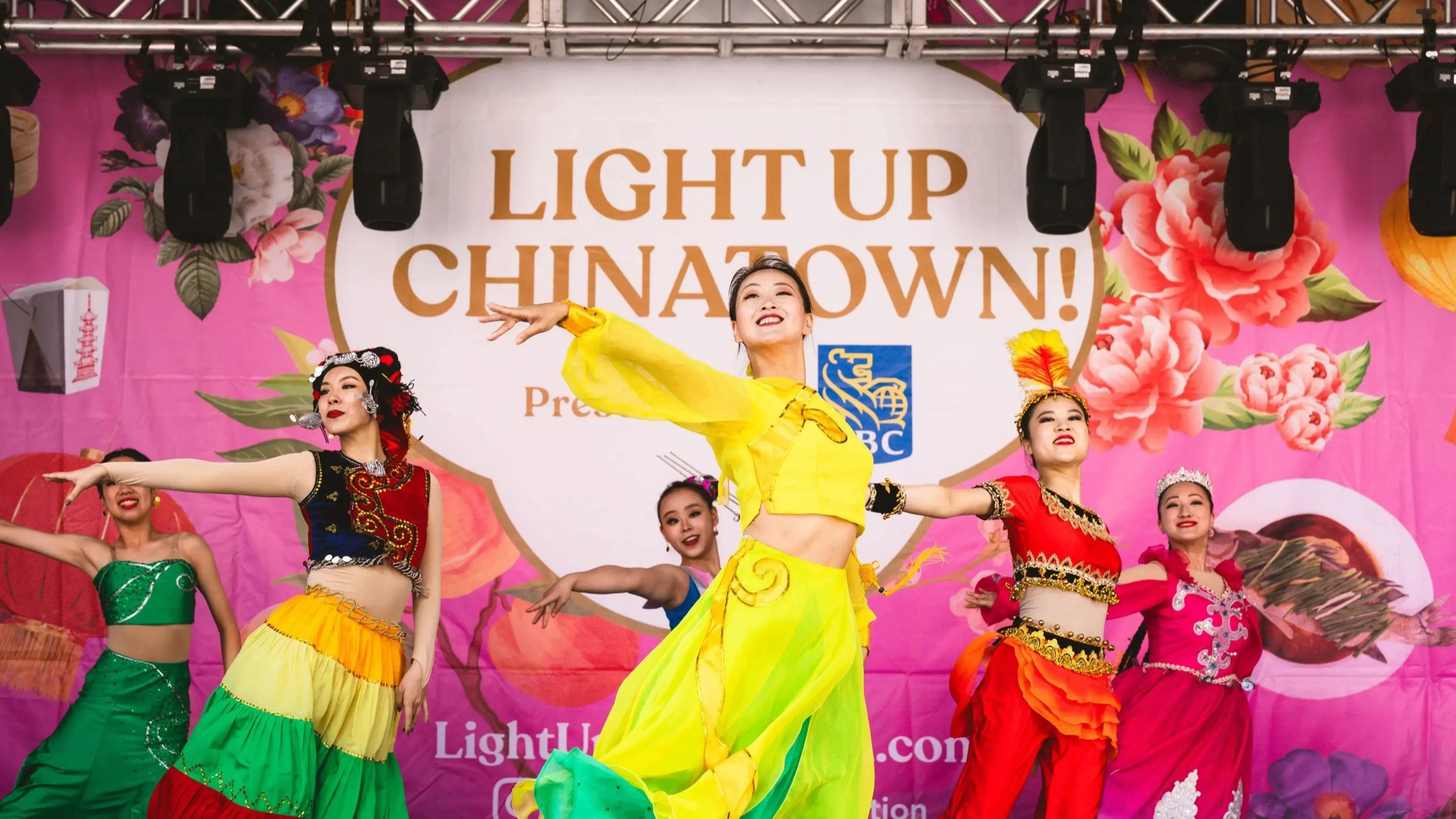 Six dancers in colorful traditional costumes performing on stage with a backdrop reading Light Up Chinatown.