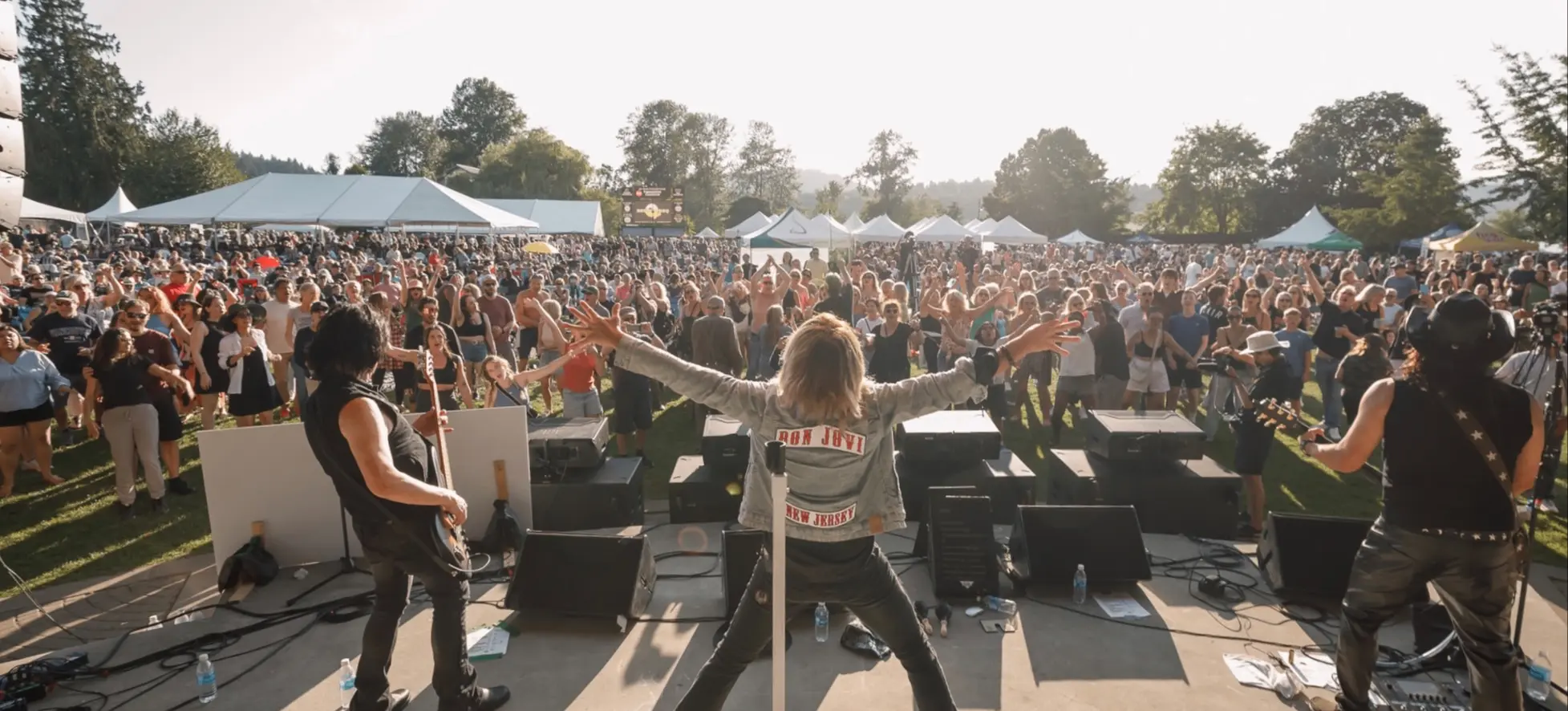 Three musicians performing on an outdoor stage facing a large crowd with tents and trees in the background.