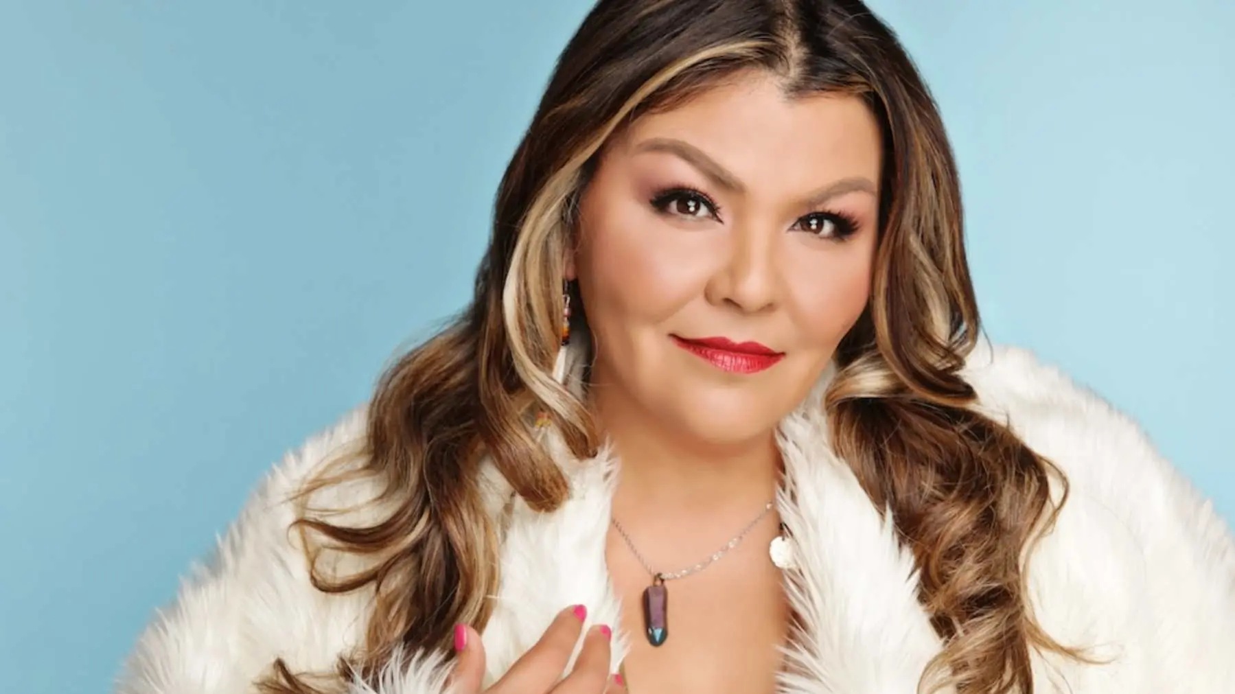 Person with long wavy hair wearing a white fur coat and a pendant necklace against a light blue background.