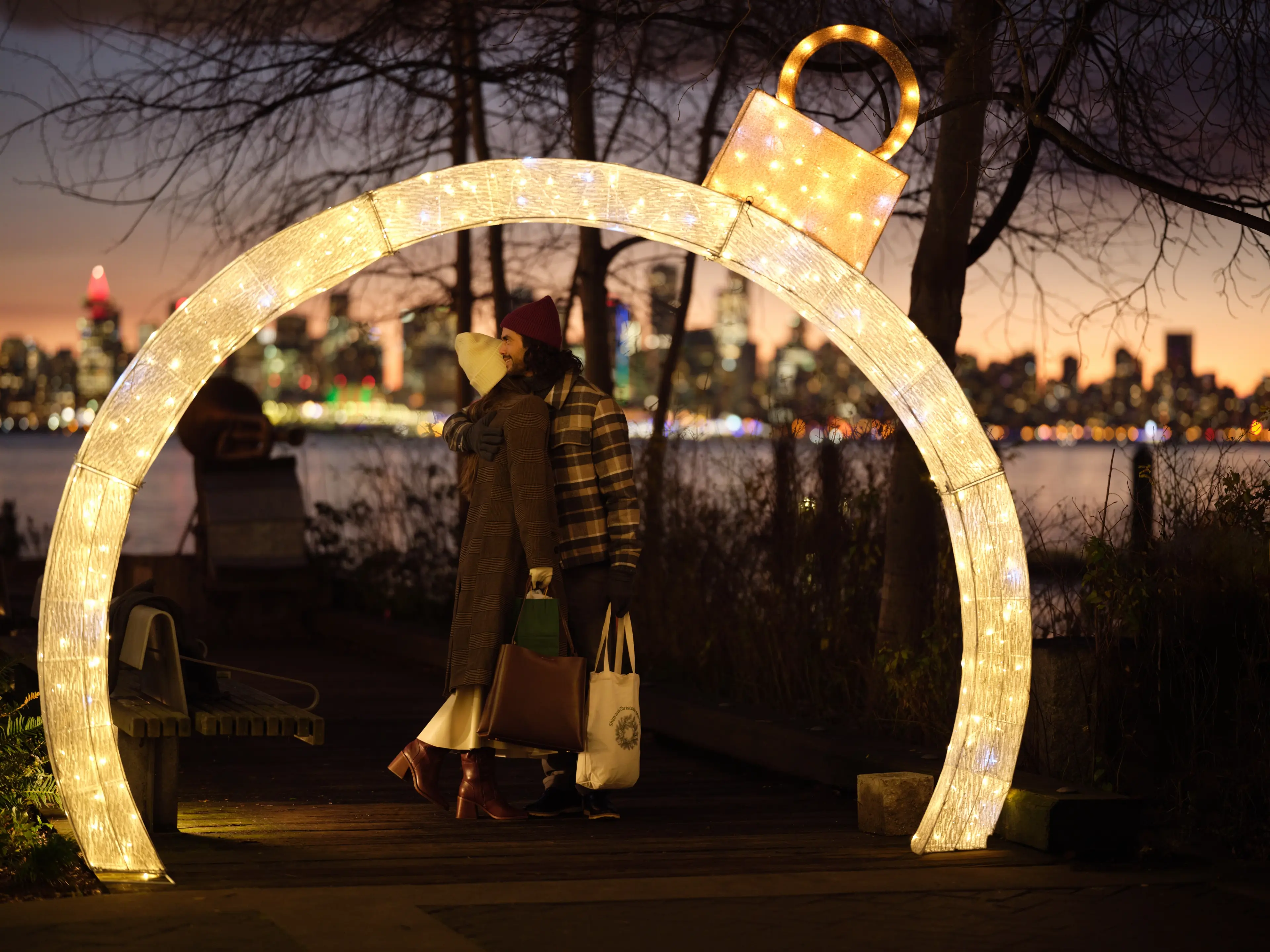 A romantic couple standing under light decorations at dusk with Vancouver downtown in the background at the North Vancouver Christmas Market