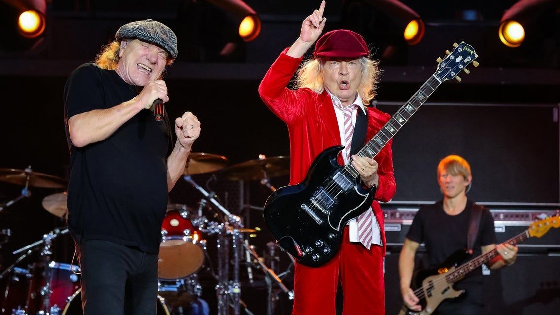 Three musicians performing on stage with one singing into a microphone, one playing electric guitar in a red suit and hat, and one playing bass guitar.