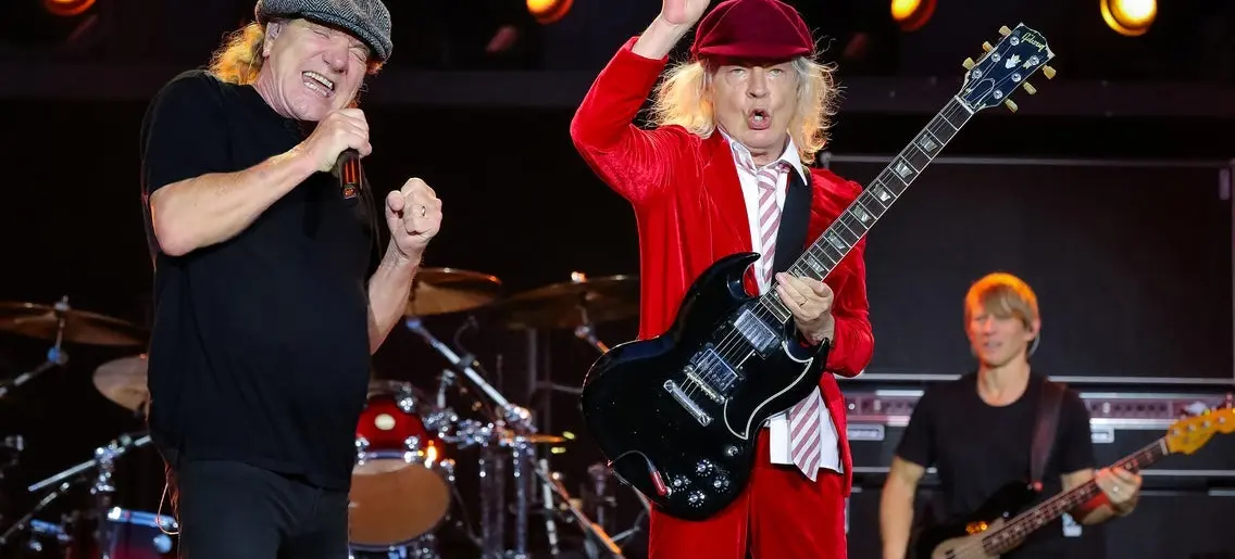 Three musicians performing on stage with one singing into a microphone, one playing electric guitar in a red suit and hat, and one playing bass guitar.