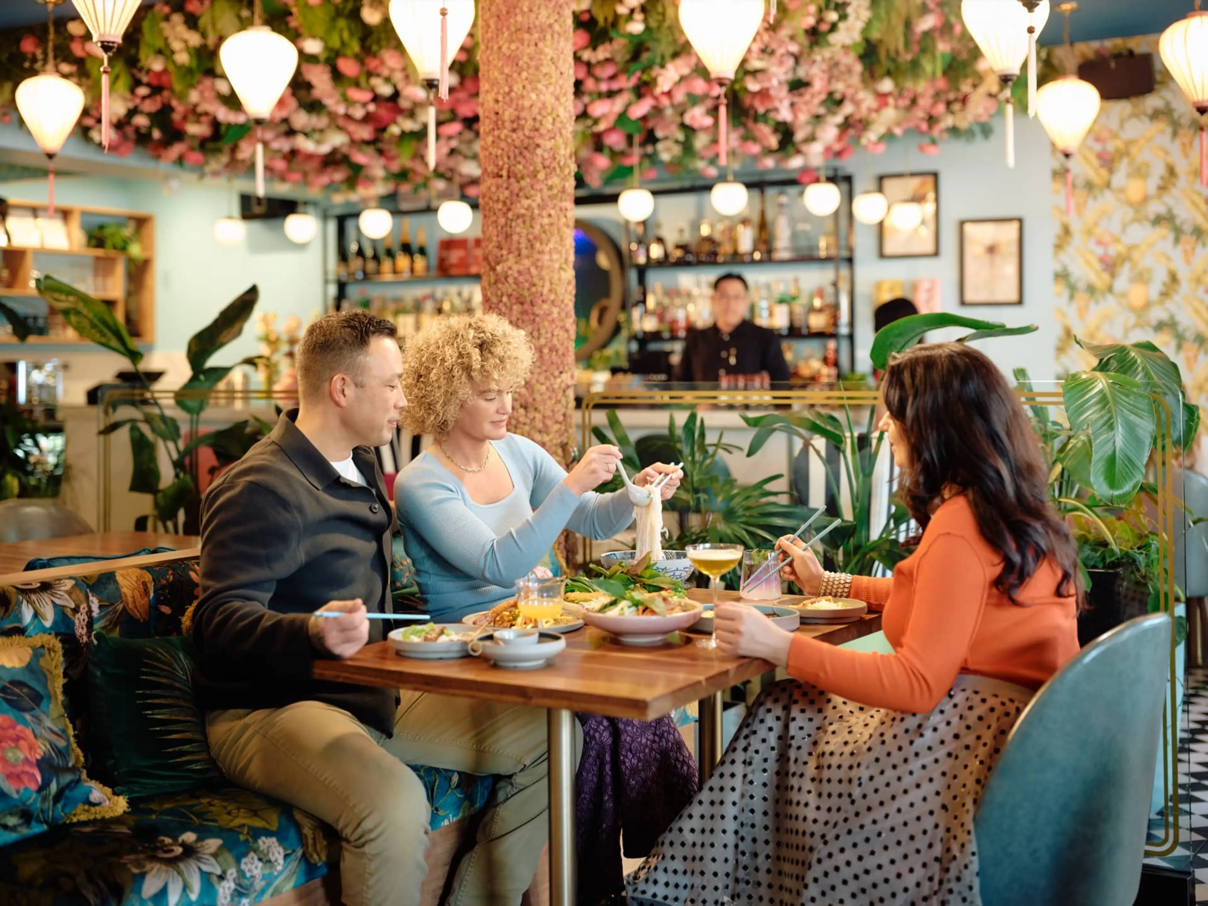 Three people sitting at a restaurant table with food, surrounded by plants and floral decor, with a bartender in the background.